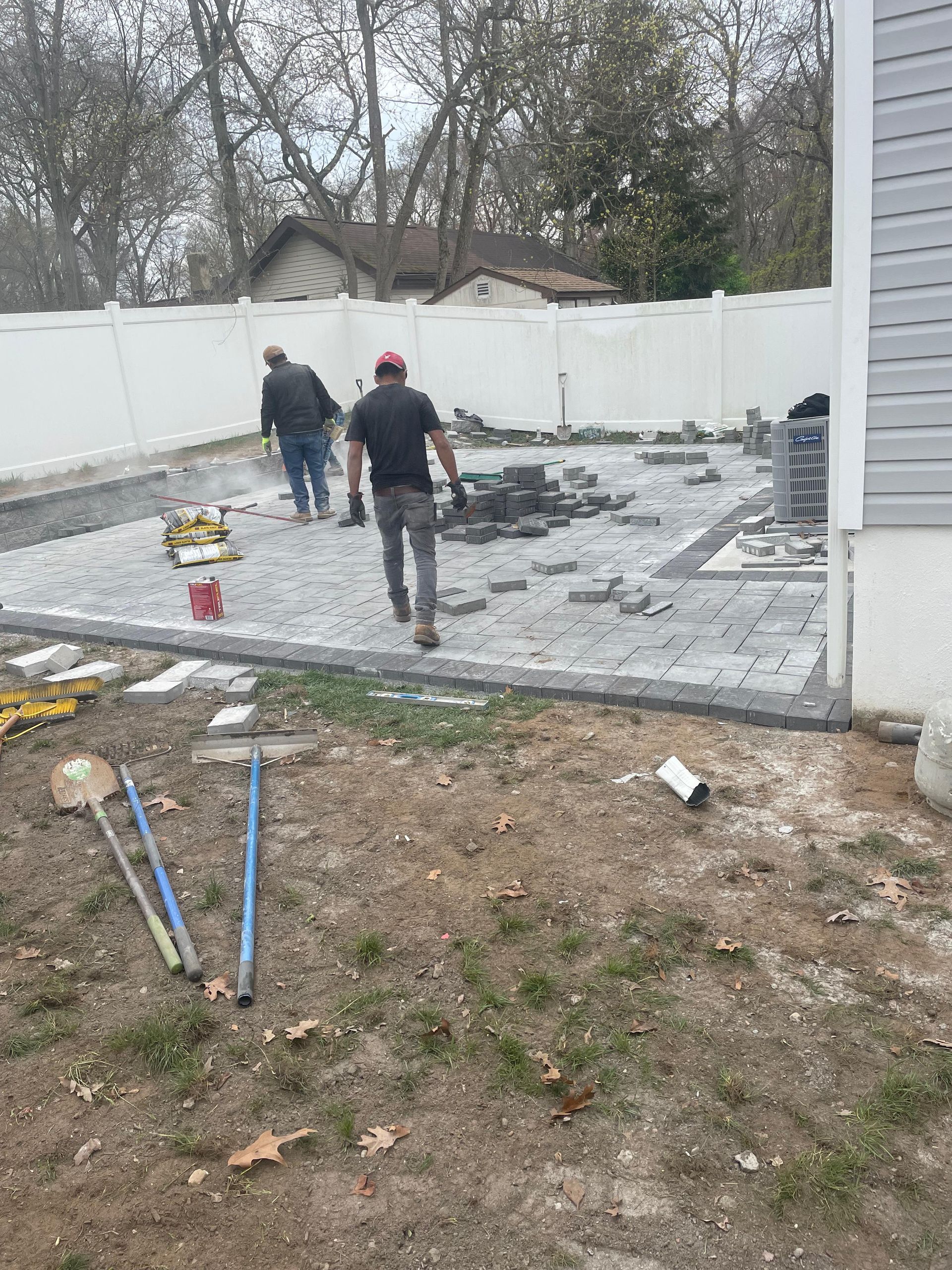 Two workers install gray stone pavers on a backyard patio enclosed by a white vinyl fence.