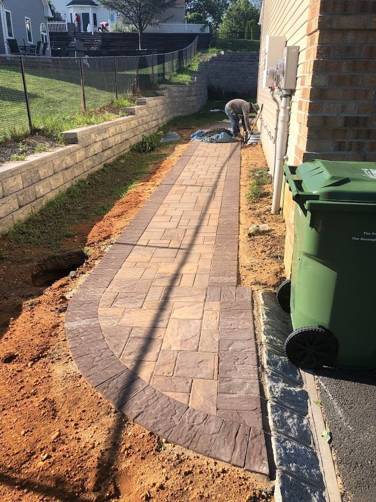 A newly installed tan and dark brown paver walkway along a brick house foundation, with a worker visible in the distance.