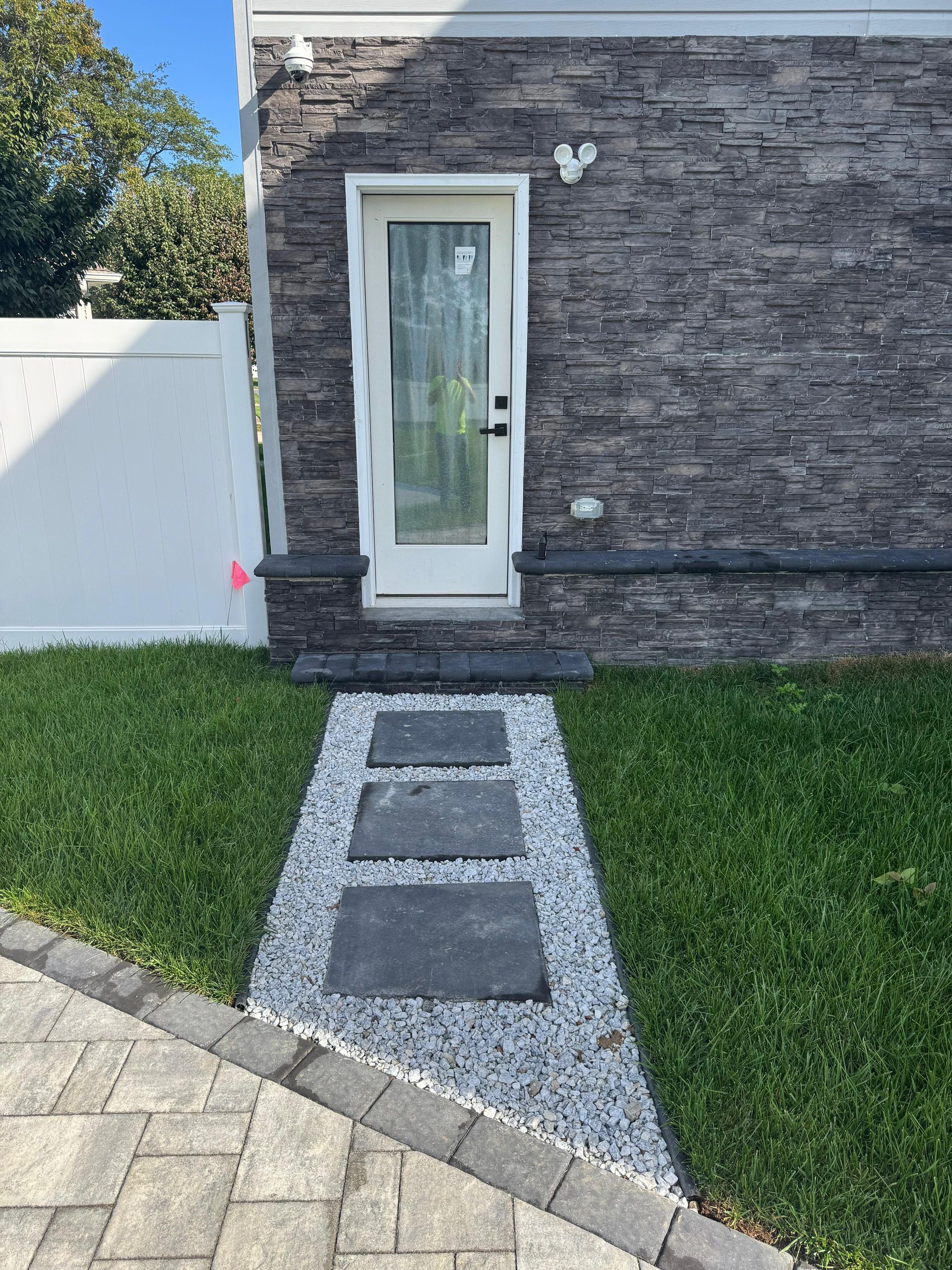 A white door set in a grey stone wall leads to a path of three square pavers surrounded by gravel through a grass yard.