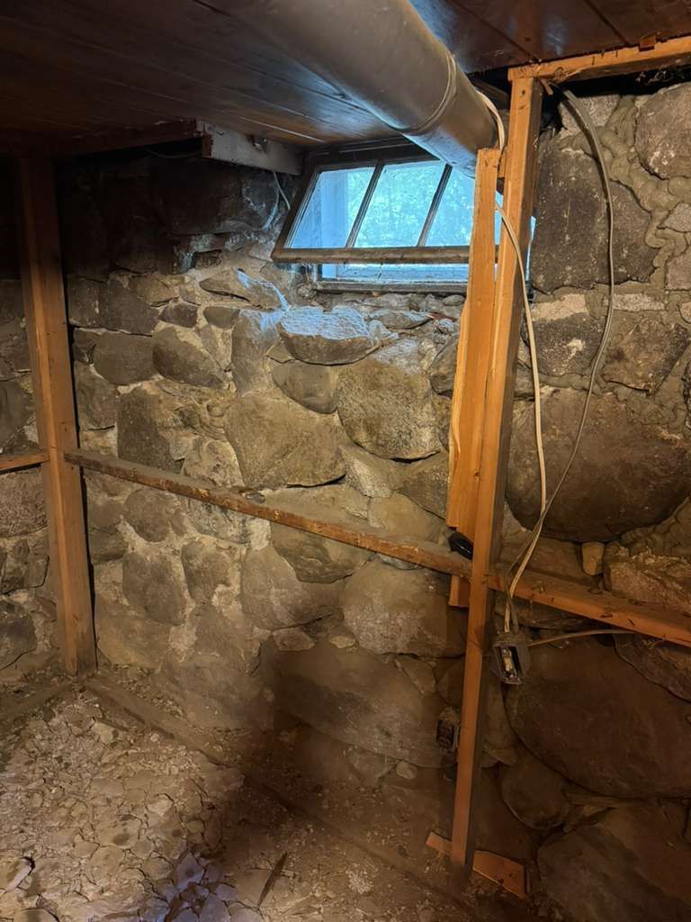 A view of a basement corner with a stone foundation wall, a small window, exposed wooden framing, and an electrical cord.