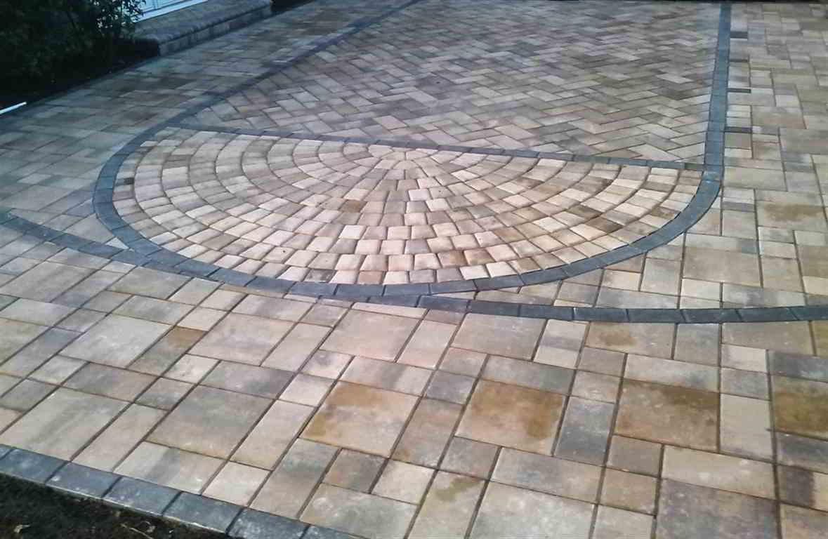 A patio paved with tan, rectangular stones, featuring a decorative fan-shaped pattern outlined by dark gray border bricks.