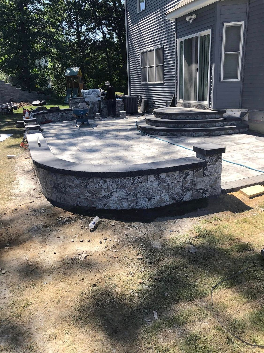 A stone patio with a curved retaining wall and steps leading to a gray house exterior in a backyard.