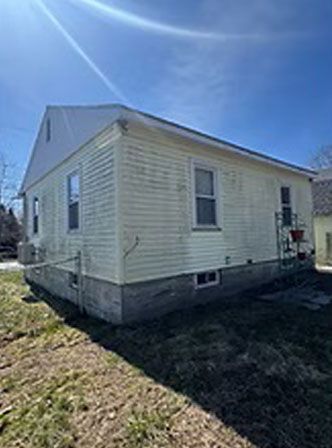 Side view of a weathered yellow house with a white roof and concrete foundation against a blue sky.