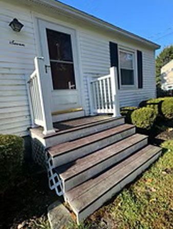 White house with wooden steps leading to the front door. Black shutters and small bushes.