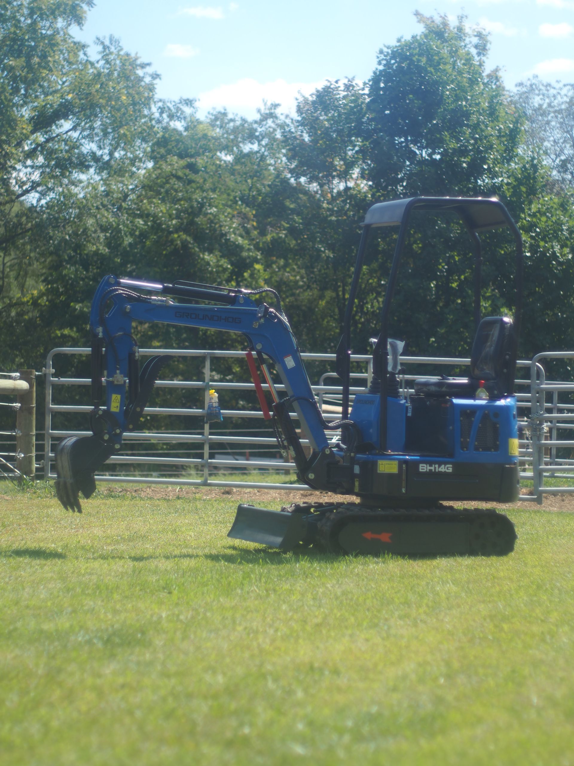 Blue mini-excavator on a grassy field with trees and a fence in the background.
