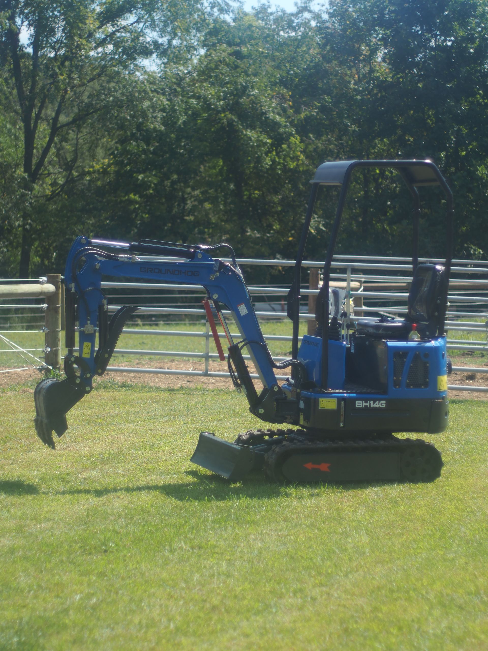 Blue mini excavator on grass, near a fence and trees.