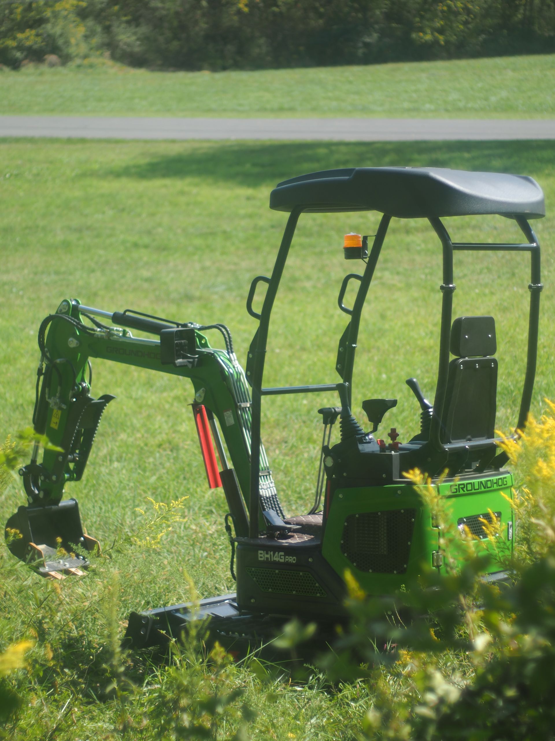 Green mini-excavator on grass with a tree line in the background.