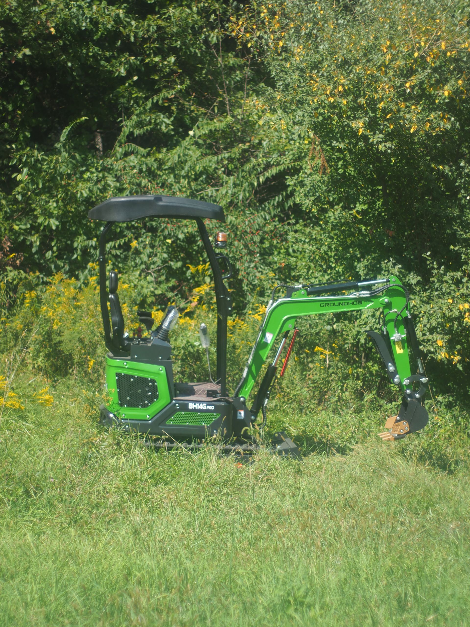 Green mini excavator in a grassy area near foliage.
