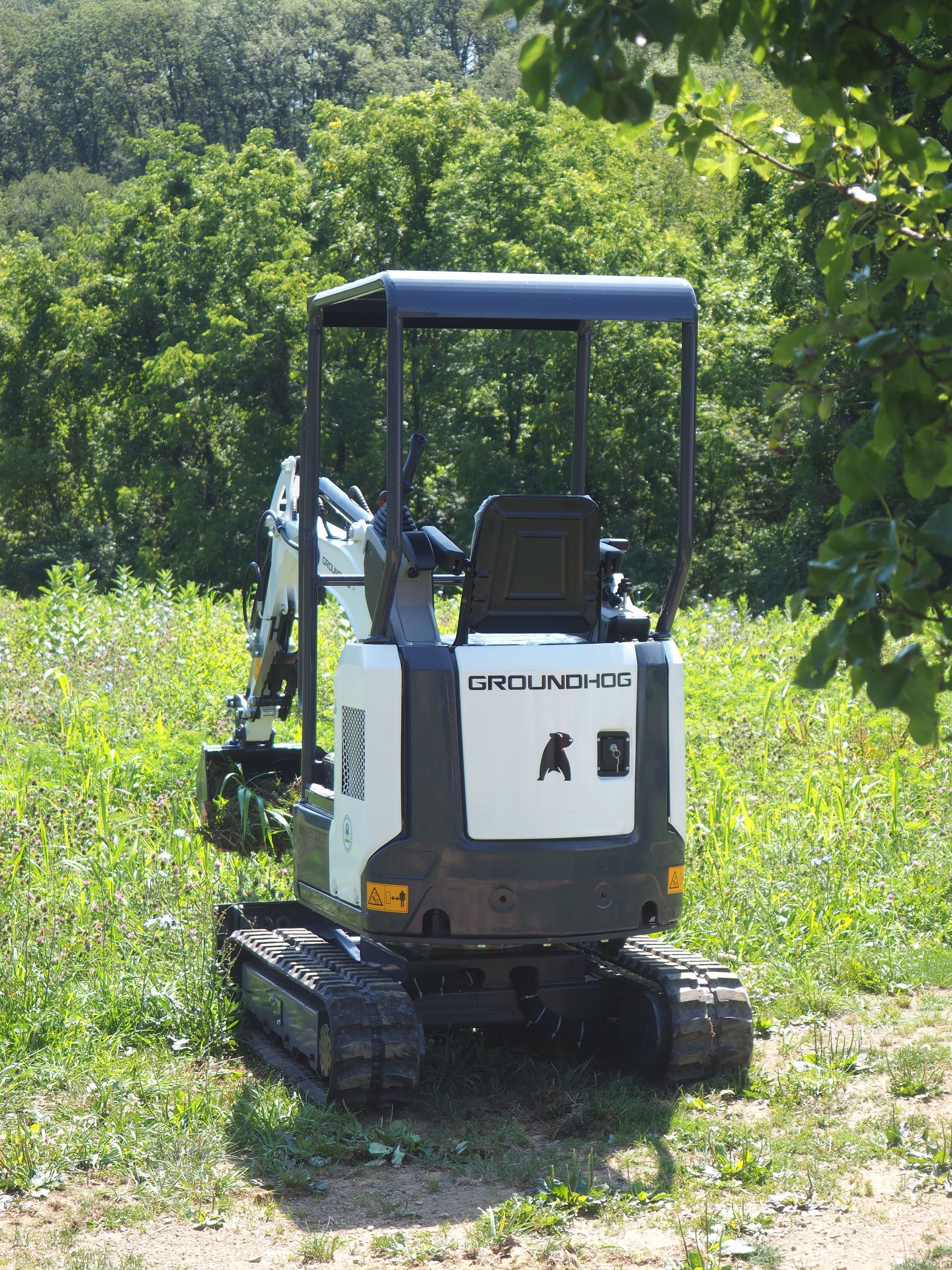 Small white and black excavator in a grassy field, with trees in the background.