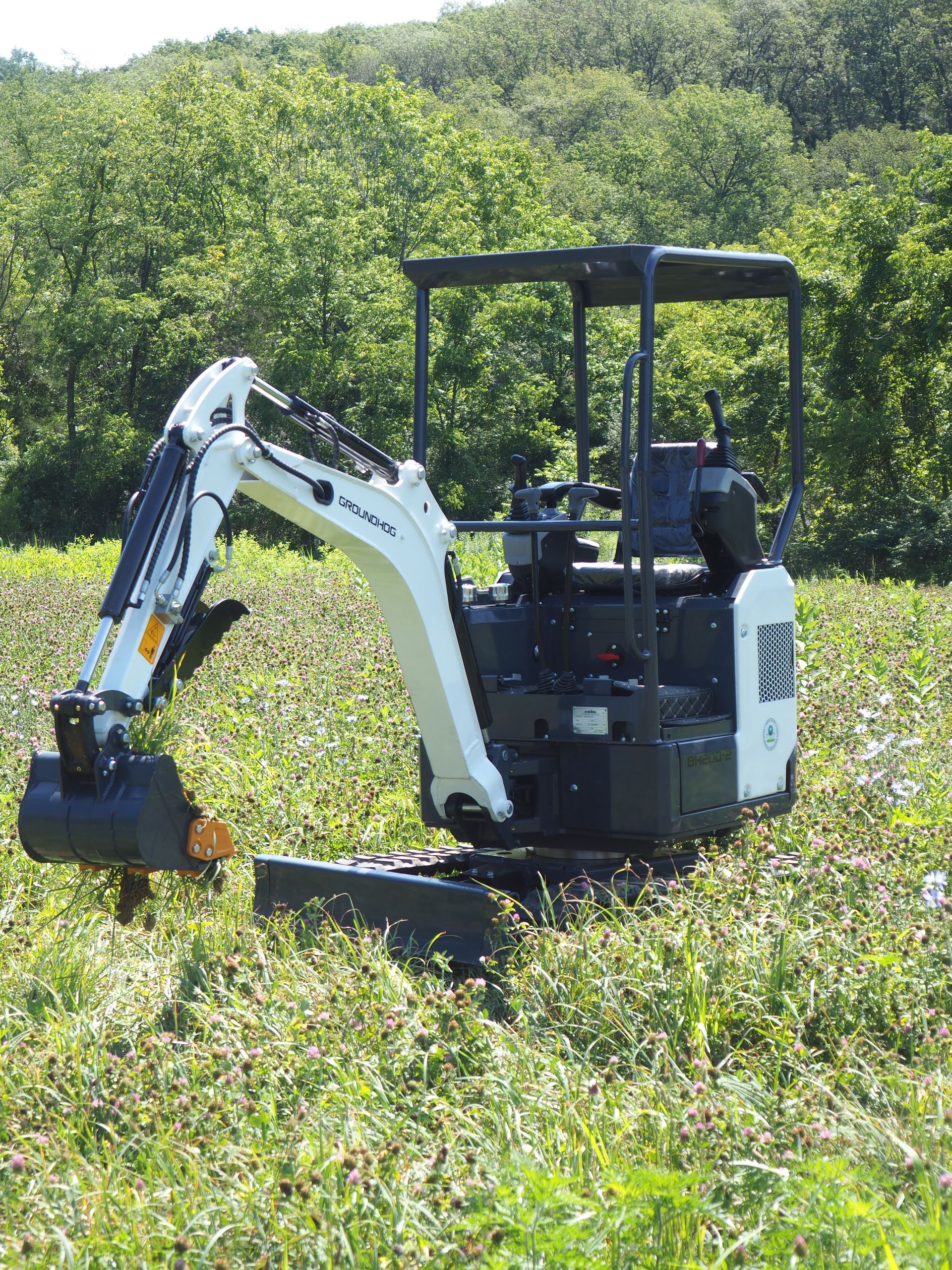 White and black mini excavator on a grassy field with trees in the background.