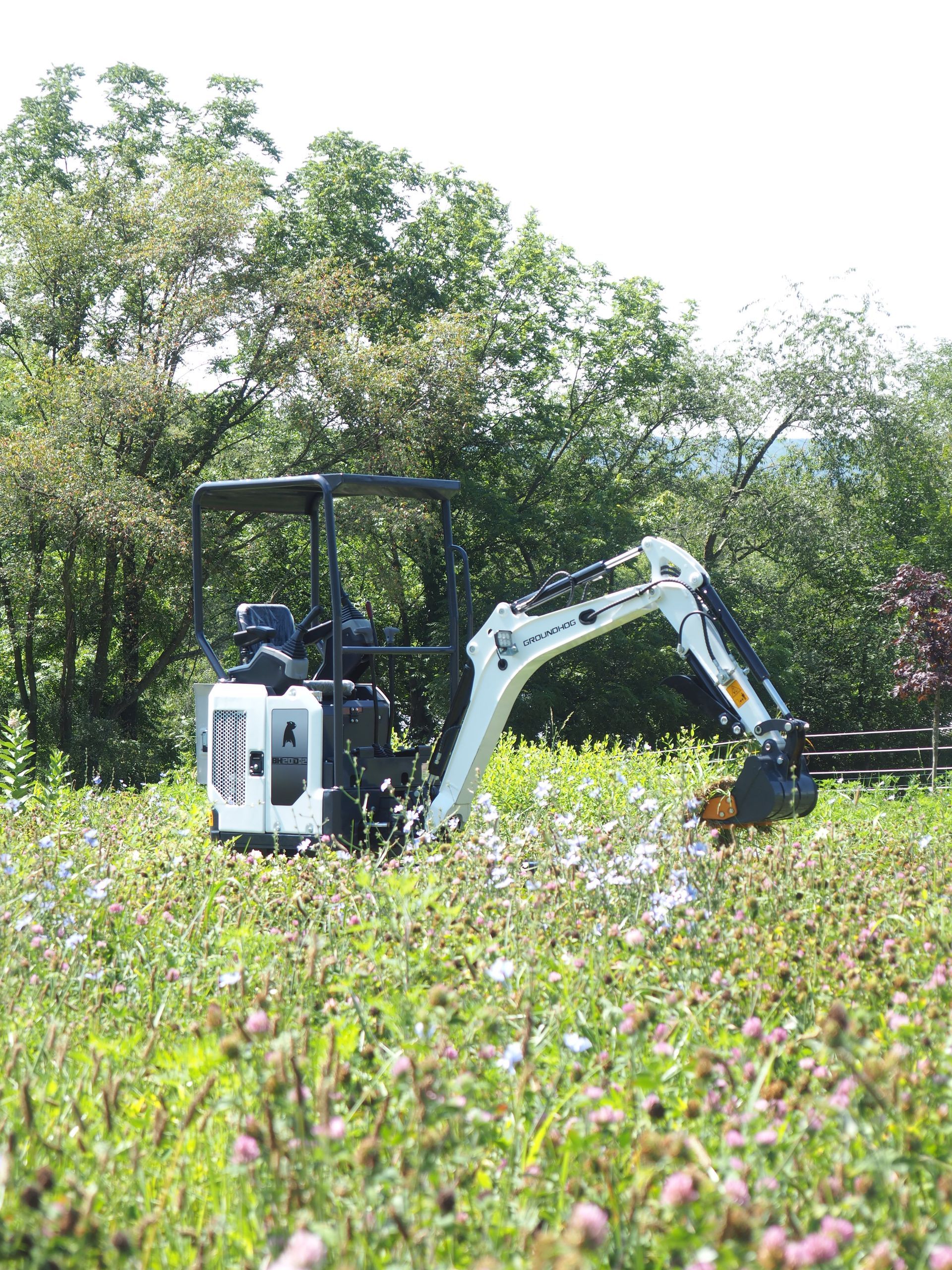 Mini excavator on a field of wildflowers, digging. Trees in the background, bright sunlight.