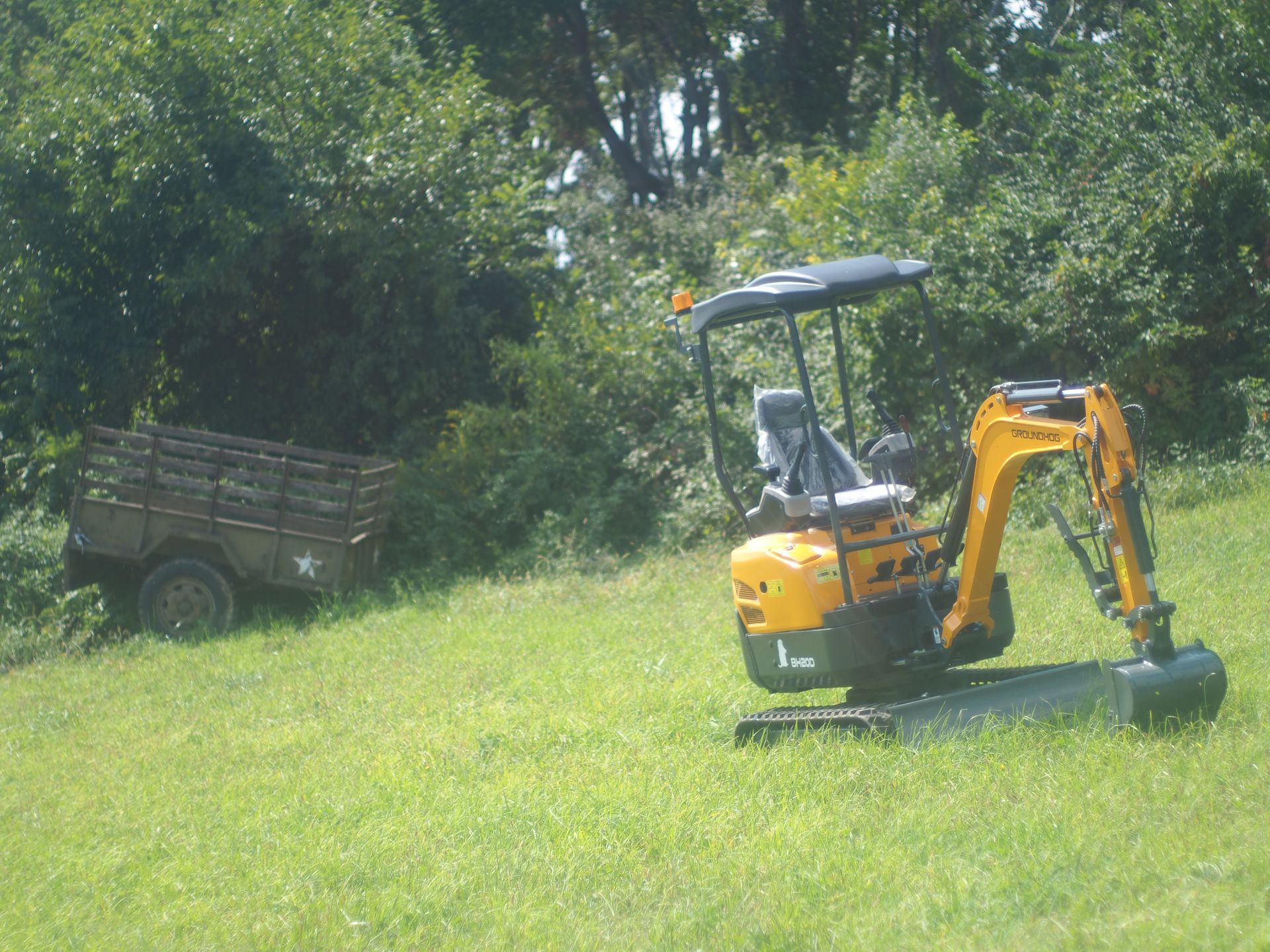 Yellow excavator on a grassy hill near a trailer and trees.