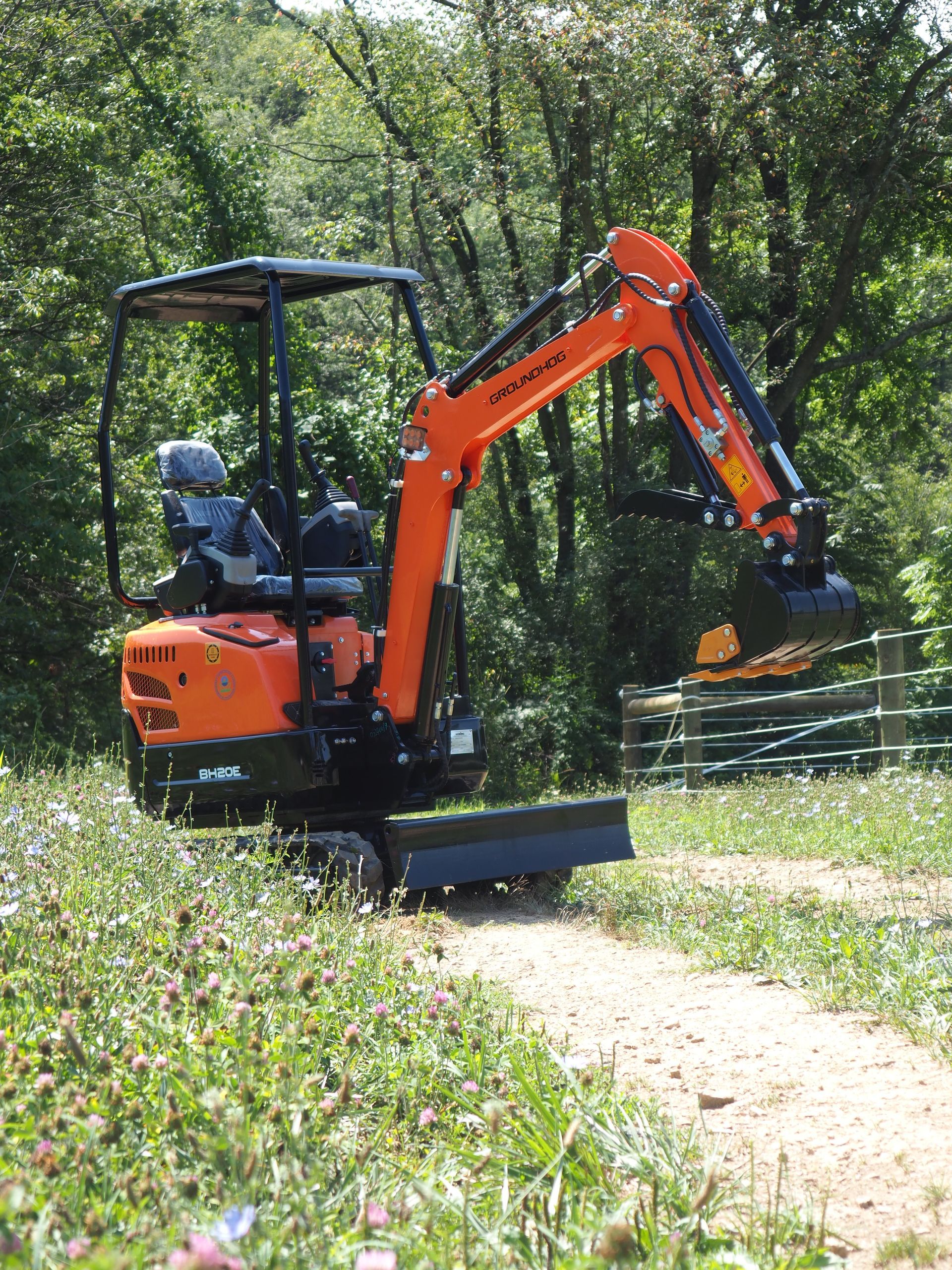 Orange mini excavator on a dirt path, poised near greenery and a wooden fence.