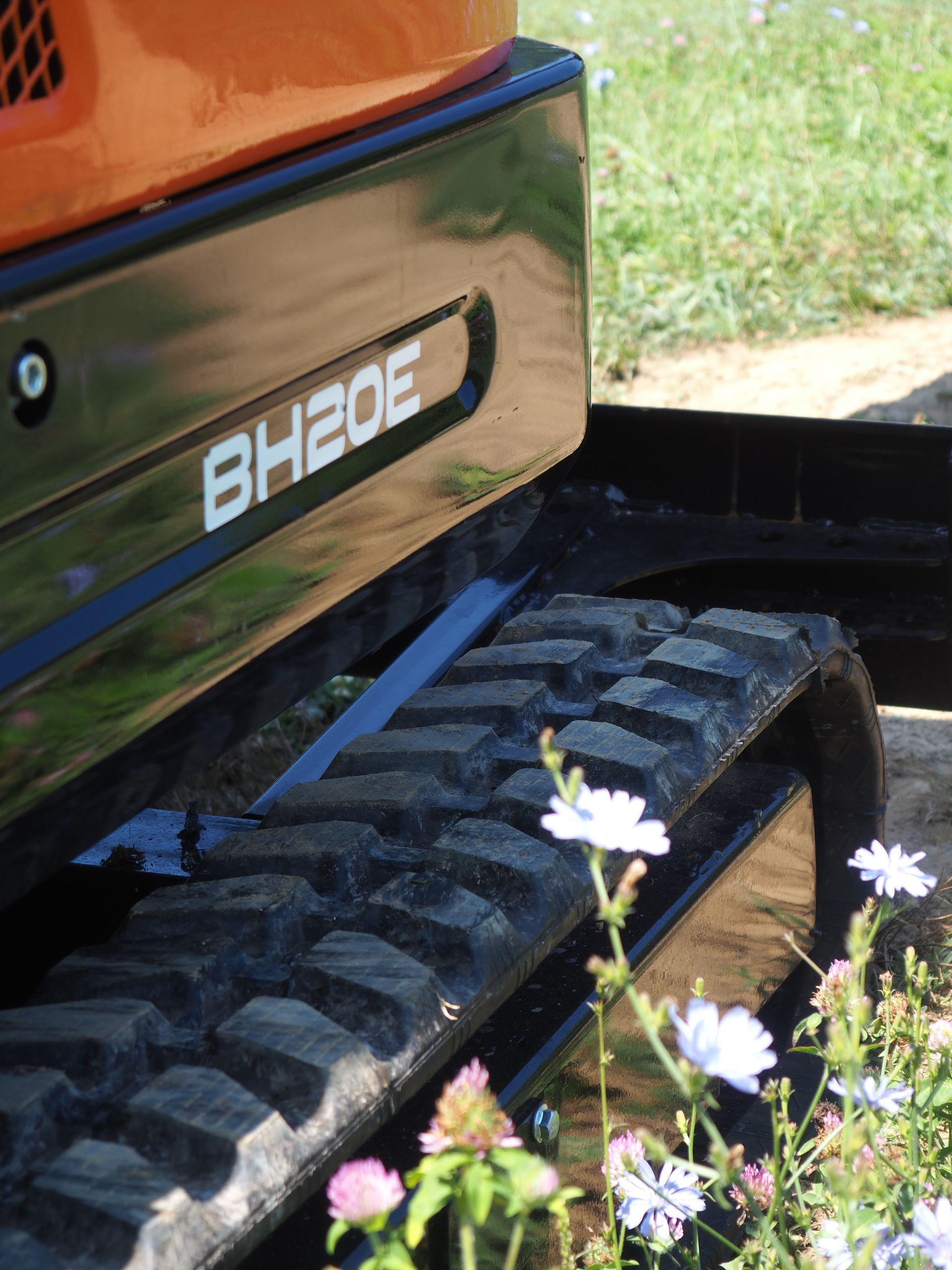 Black and orange mini excavator labeled BH20E with track and wildflowers in foreground.