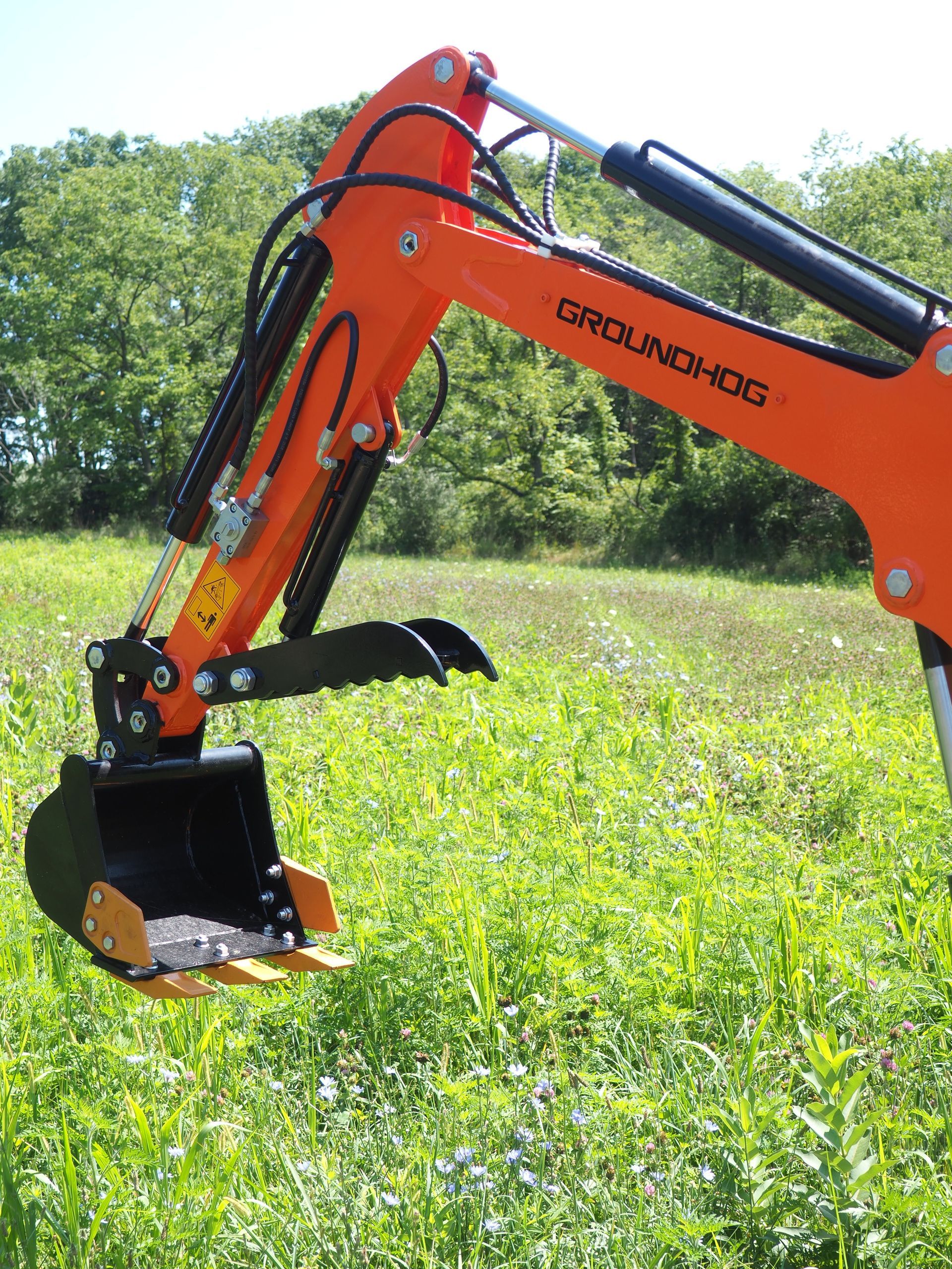 Orange Groundhog backhoe arm with bucket in a grassy field.