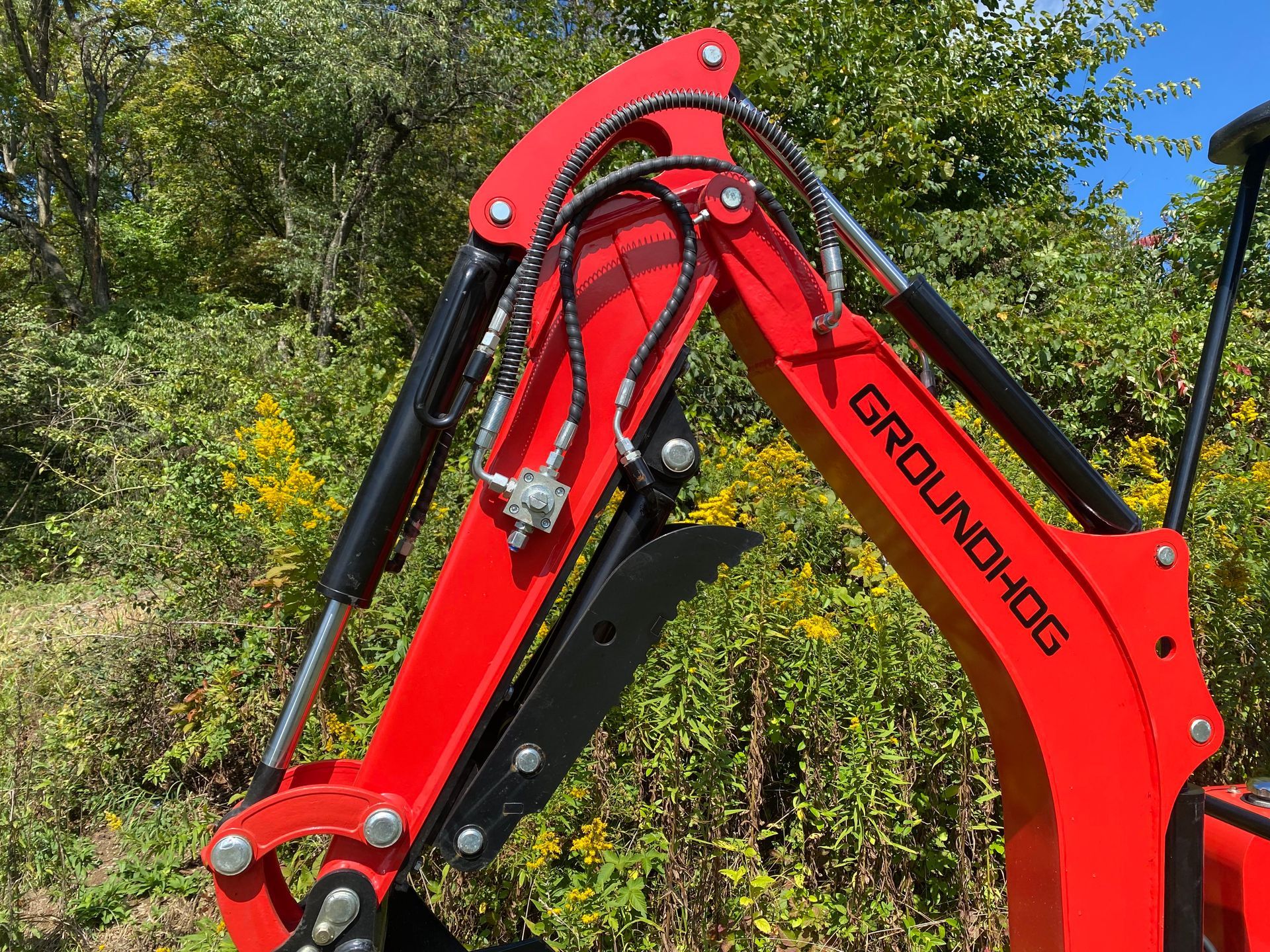 Red Groundhog mini-excavator arm, hydraulics visible, outdoors with foliage in background.