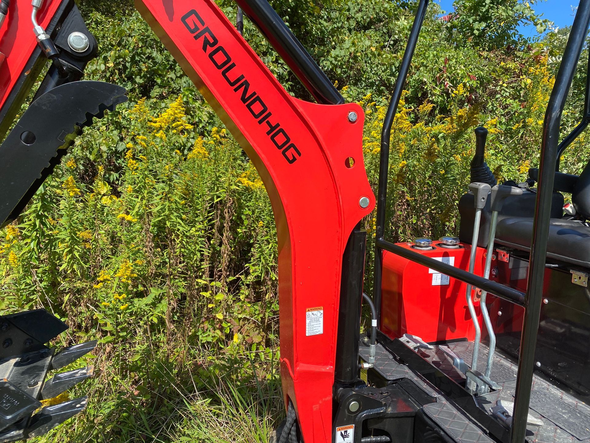 Red GROUNDHOG excavator arm against a backdrop of green foliage.