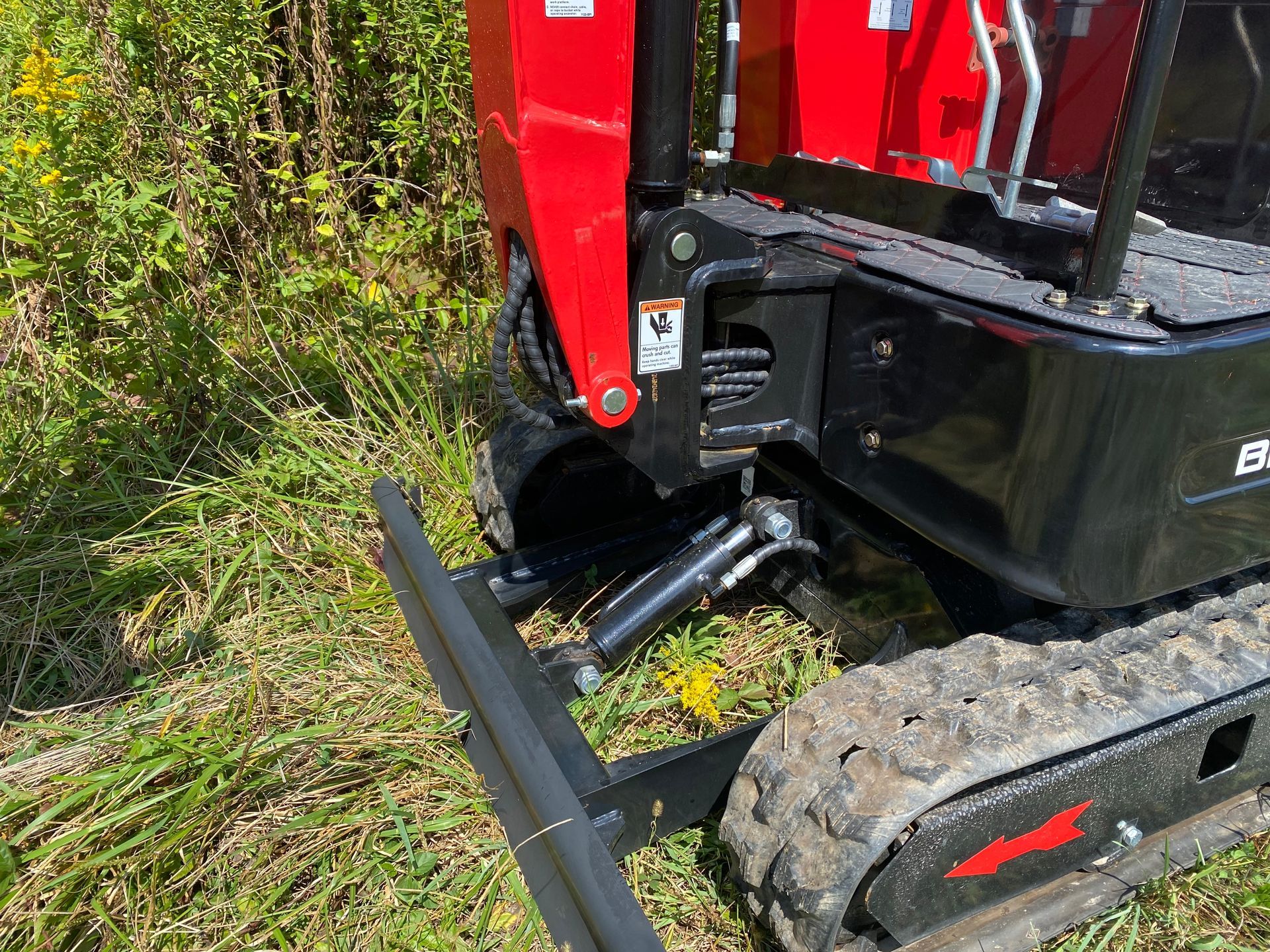 Red and black mini excavator on grass; tracks, arm, and stabilizer visible.