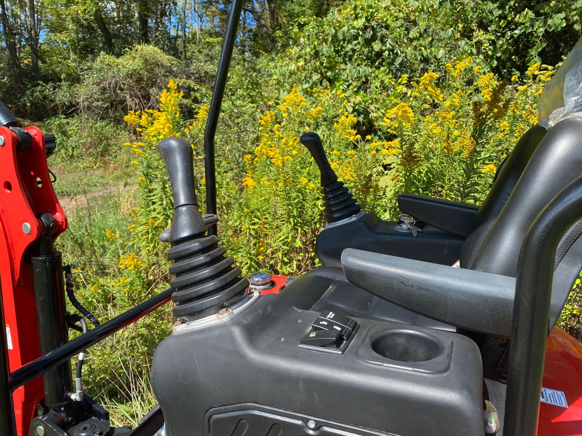Interior view of an excavator's cab with black controls and a cup holder, with a yellow flower field in the background.