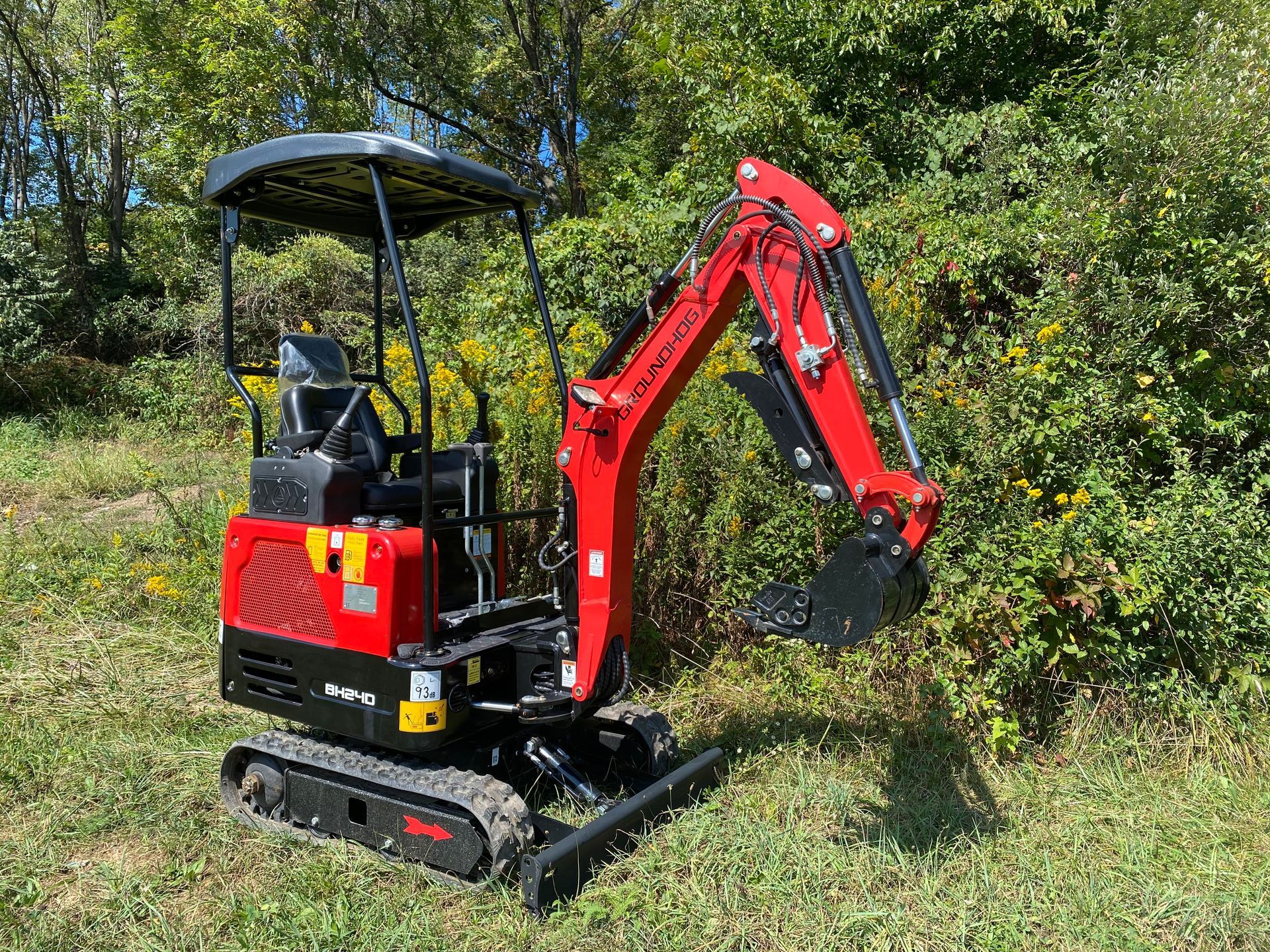 Red and black mini-excavator in grassy field. Sun shining, arm raised, ready to dig.