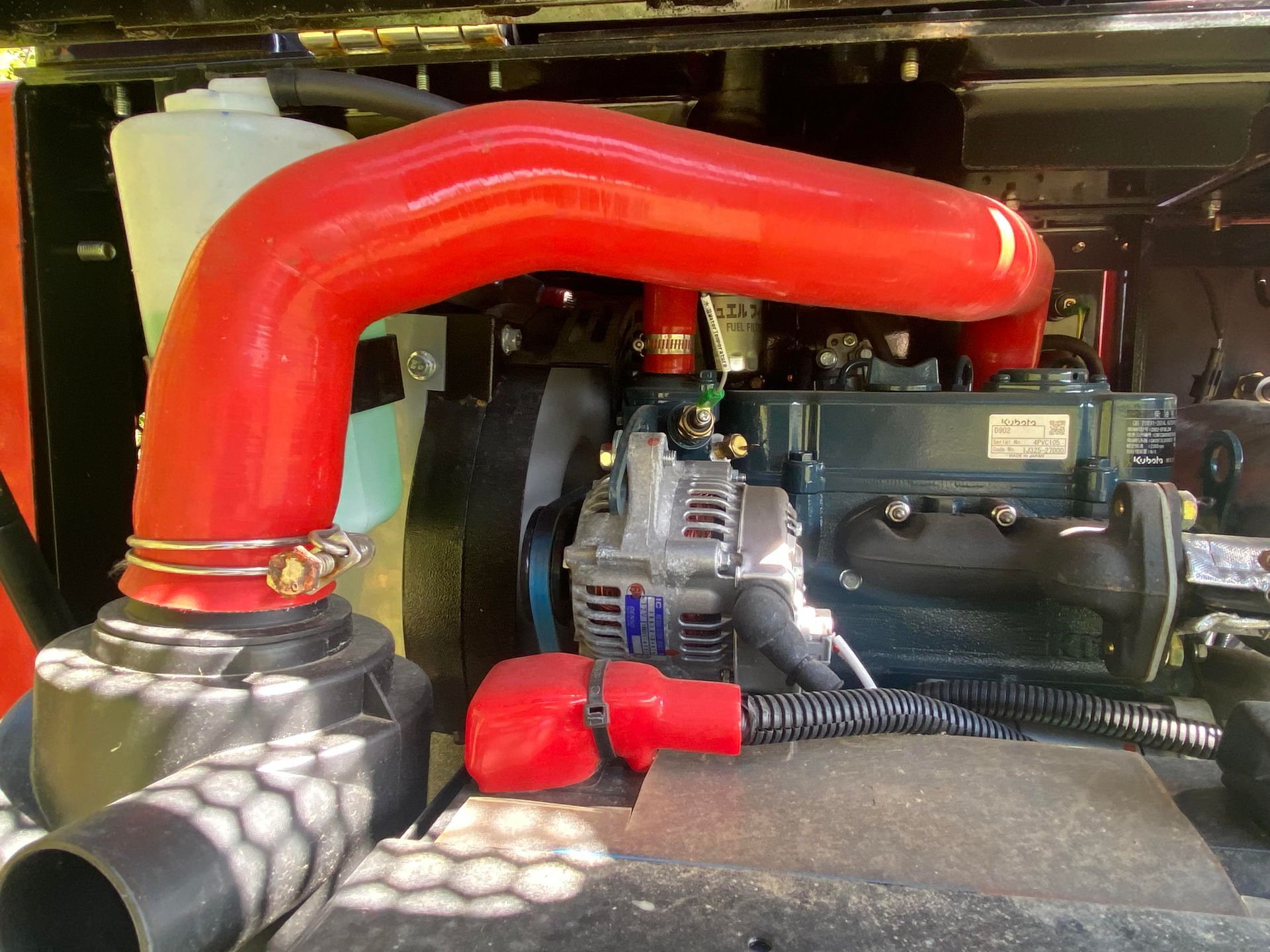 Red engine hose curves above a dark engine with visible alternator and red positive battery terminal.