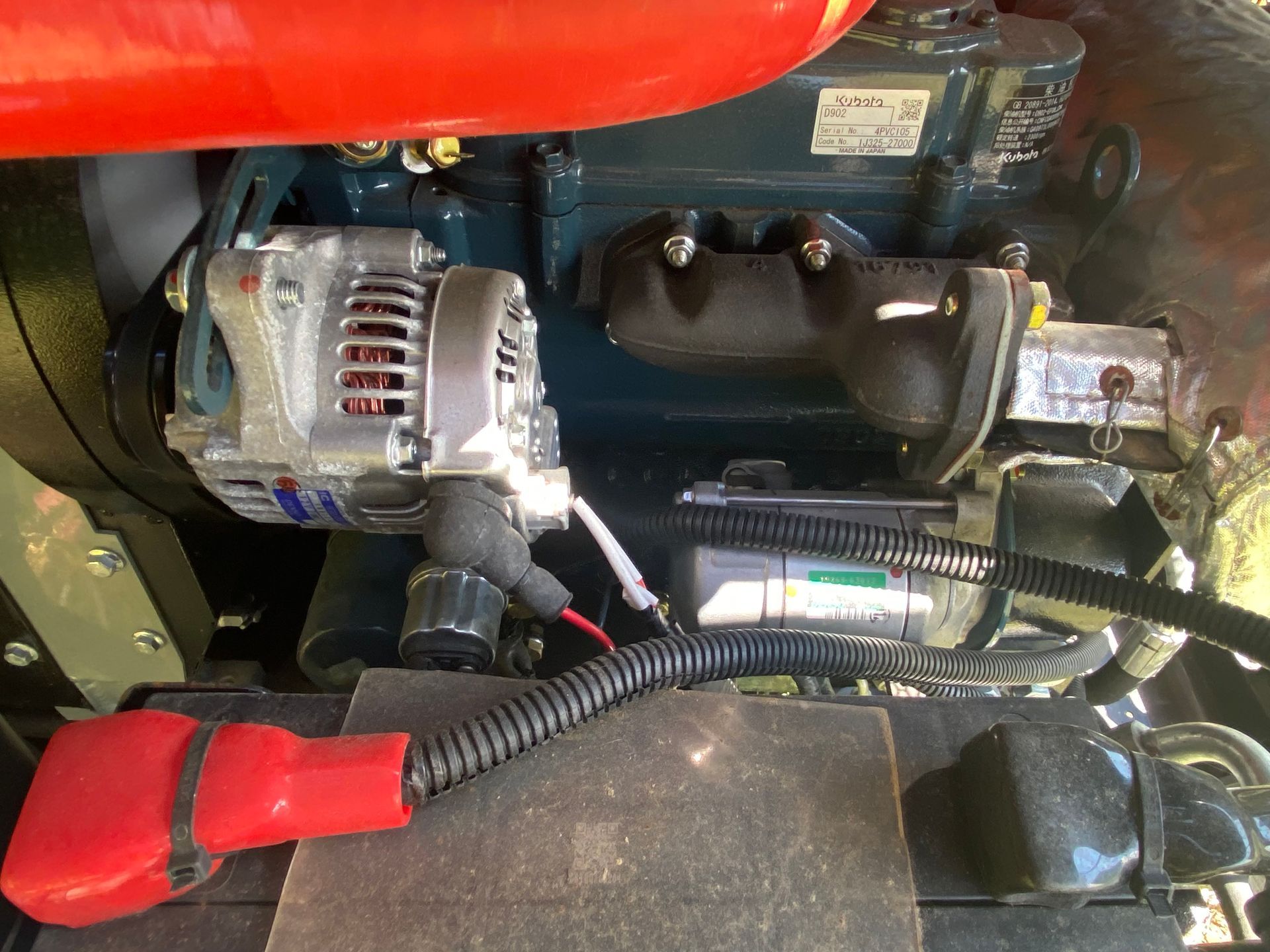 Engine components, including an alternator, on a machine with a red cable connected to a battery.