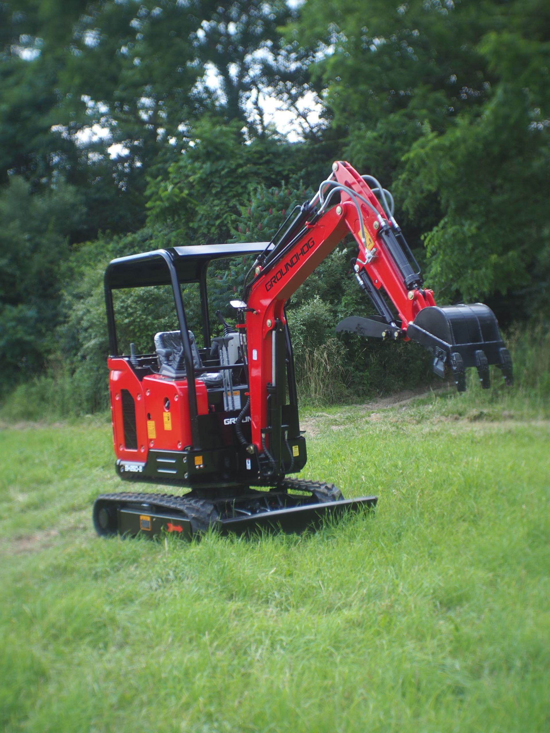 Red and black mini excavator on grass, arm extended, in a field with trees in the background.
