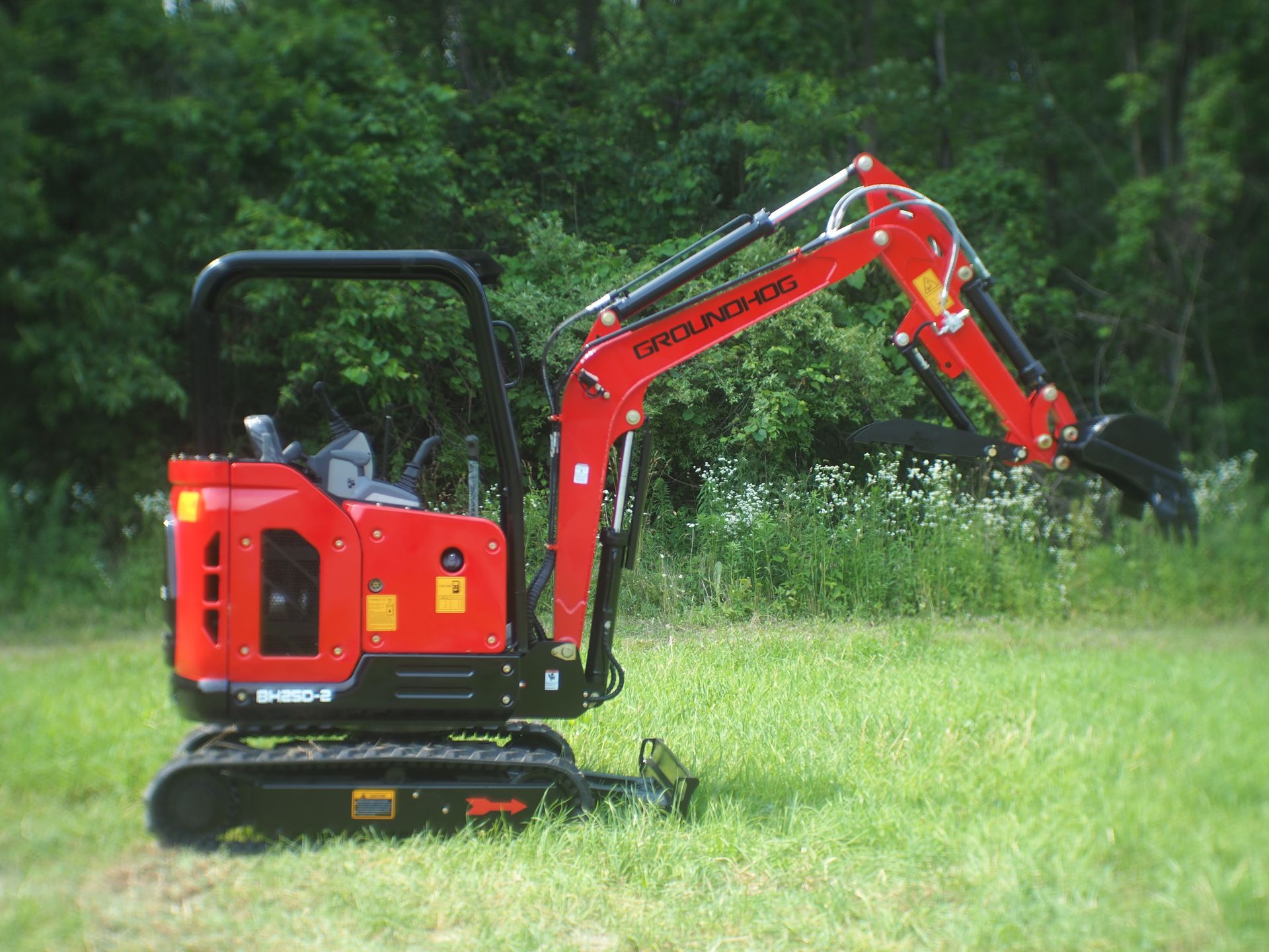Red mini excavator on grass, digging, in front of a forest.