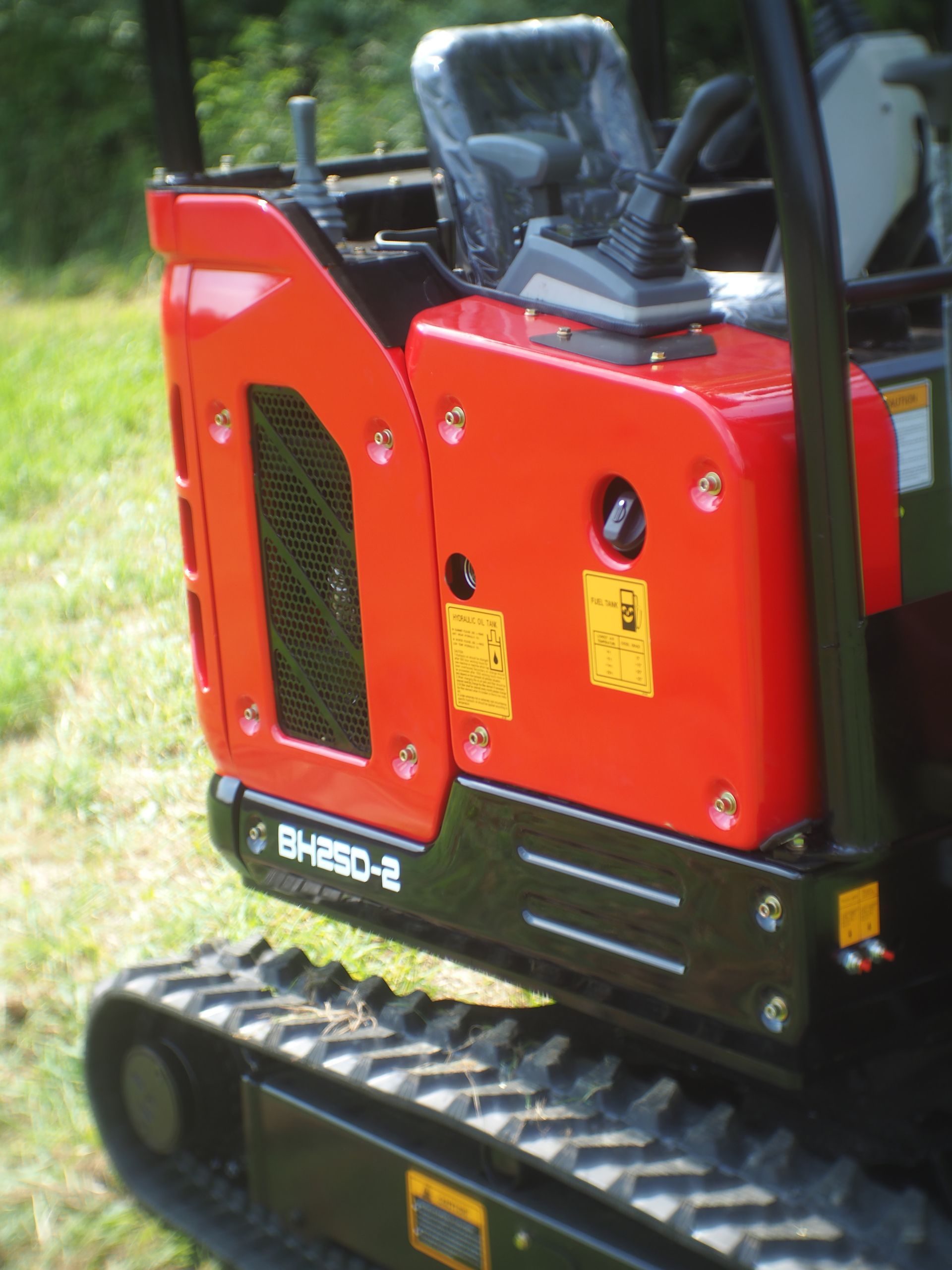 Red and black mini-excavator, outdoors. Side view showing tracks, engine cover, and control panel.