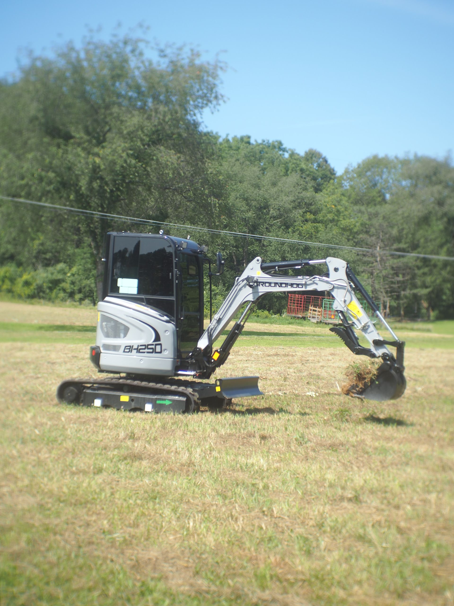Small excavator on a grassy field, scooping up dirt, under a blue sky.