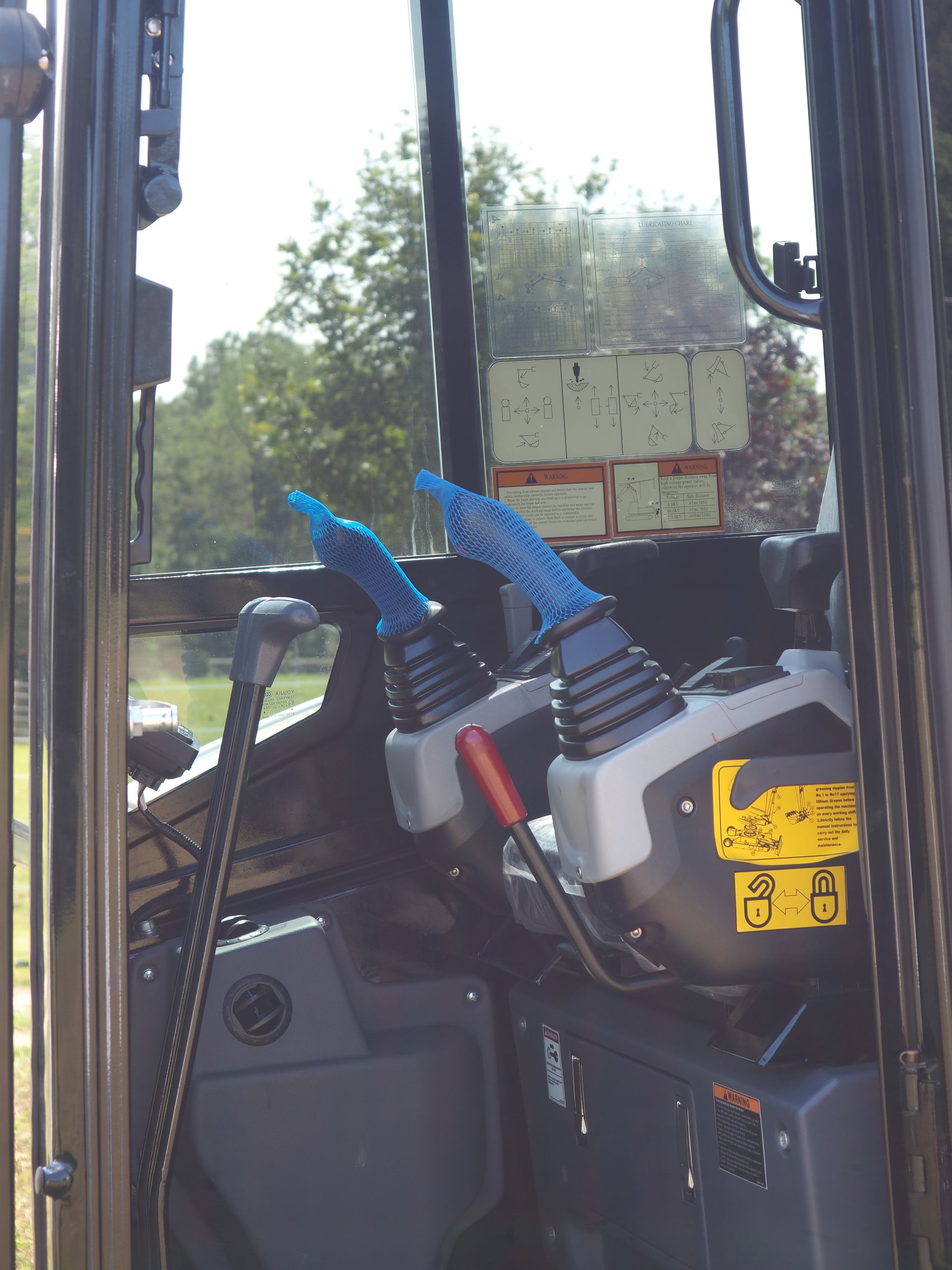 Interior view of a construction vehicle. Two joysticks with blue covers, a red lever, and yellow warning labels are visible.