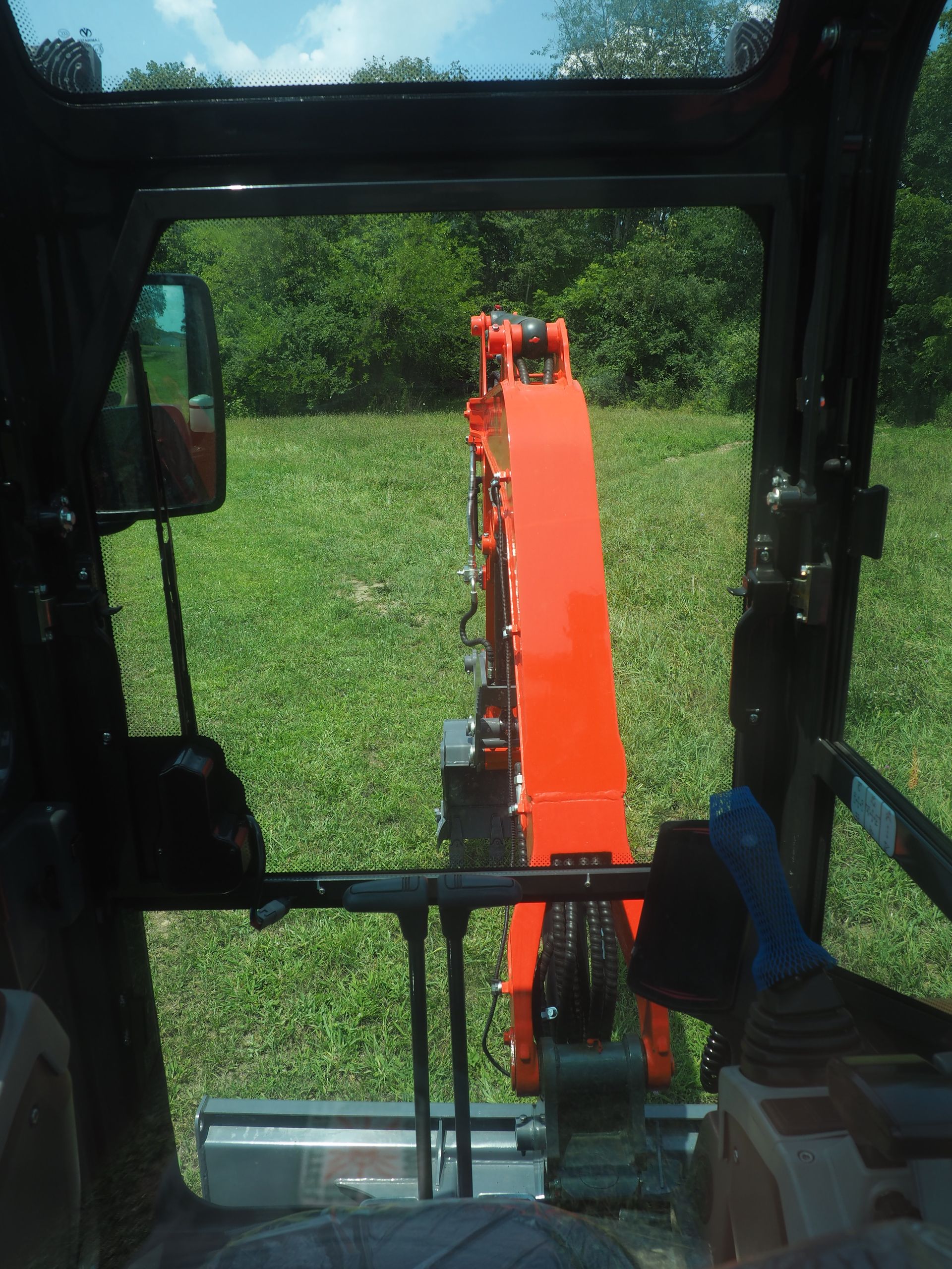 View from inside an excavator cab. Orange arm and bucket in grassy field.