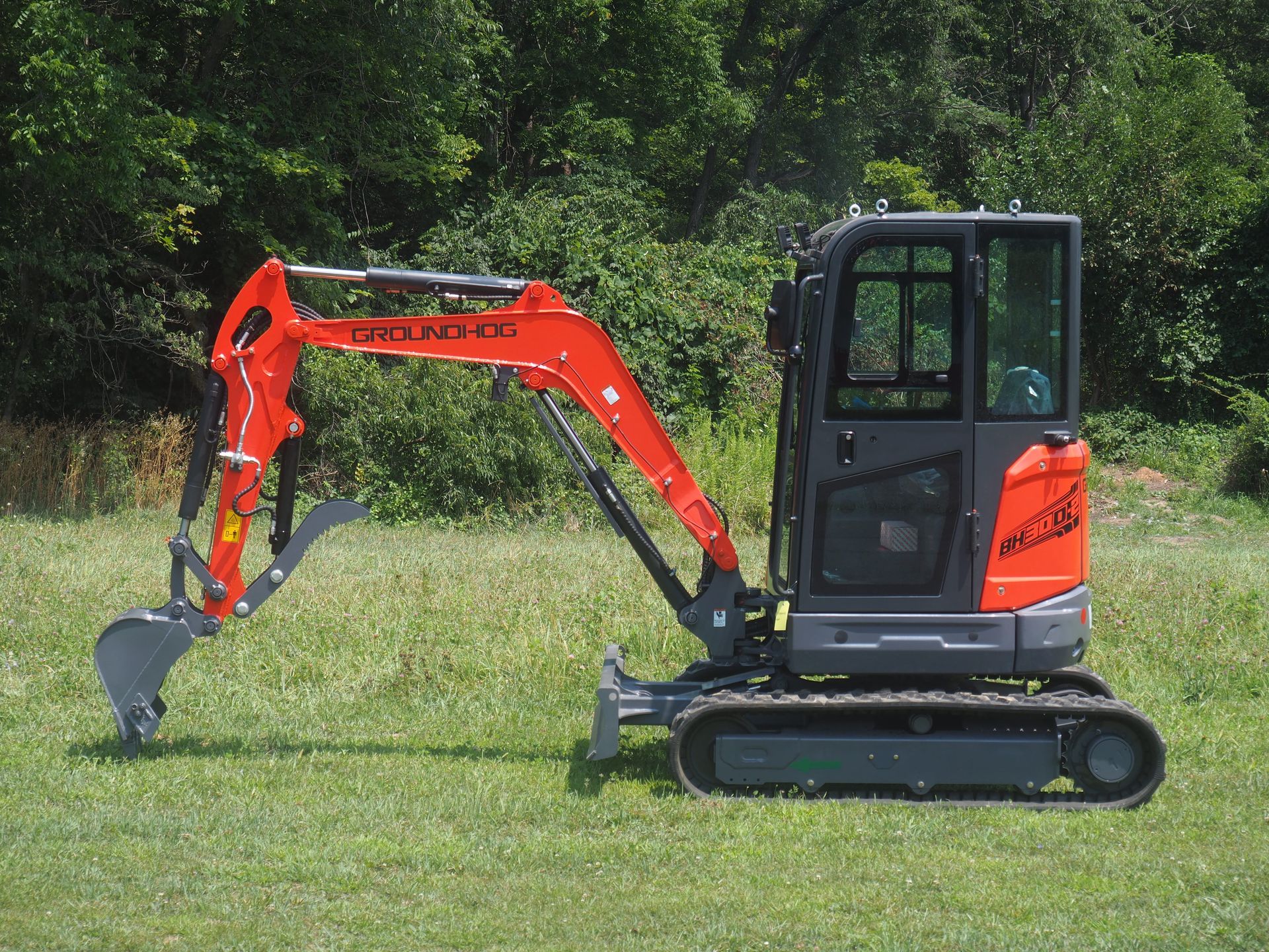 Orange and gray mini excavator on a grassy field, with a forest backdrop.