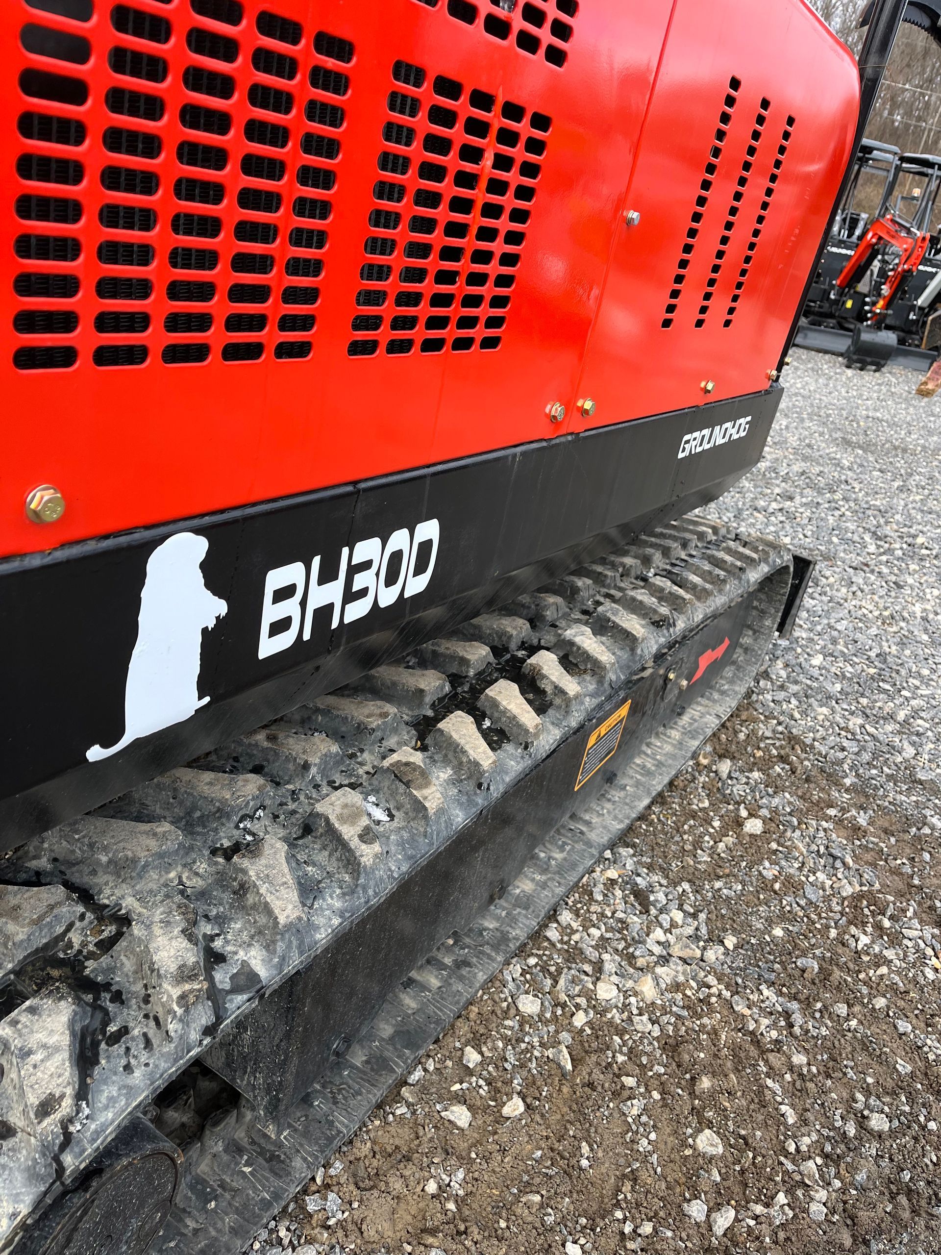 Close-up of a red and black mini-excavator with BH300 and a logo on the side, next to a muddy track.