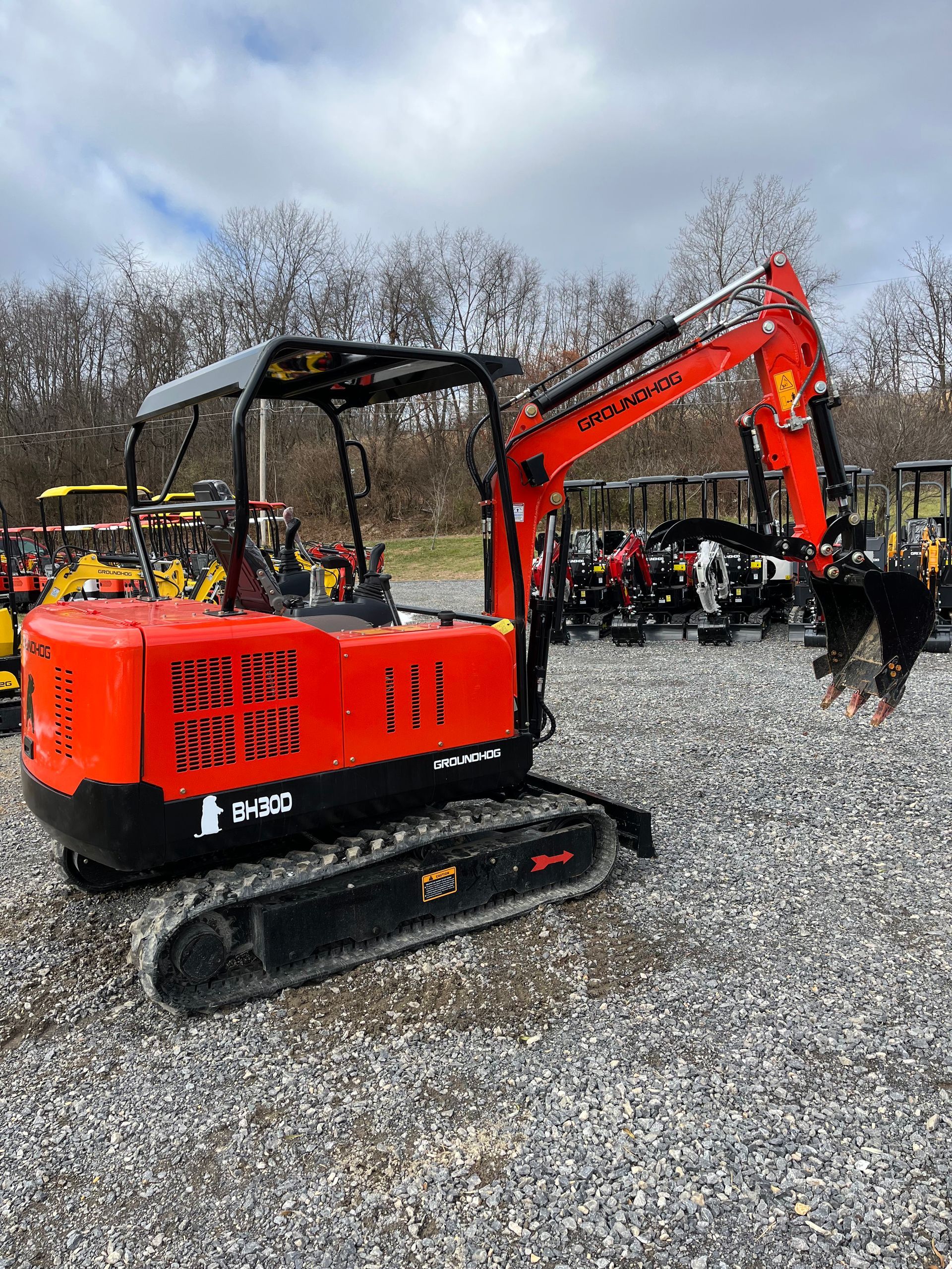 Orange mini-excavator on gravel lot, arm extended with bucket, trees in background.