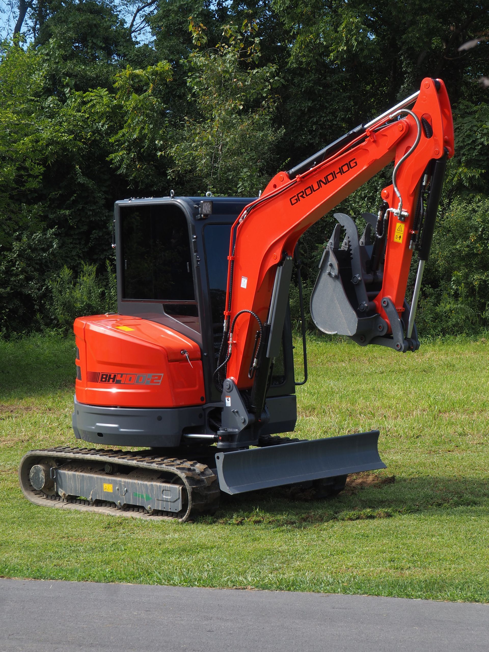 Red Kubota mini excavator on grass, with bucket, blade, and dark cab; trees in background.