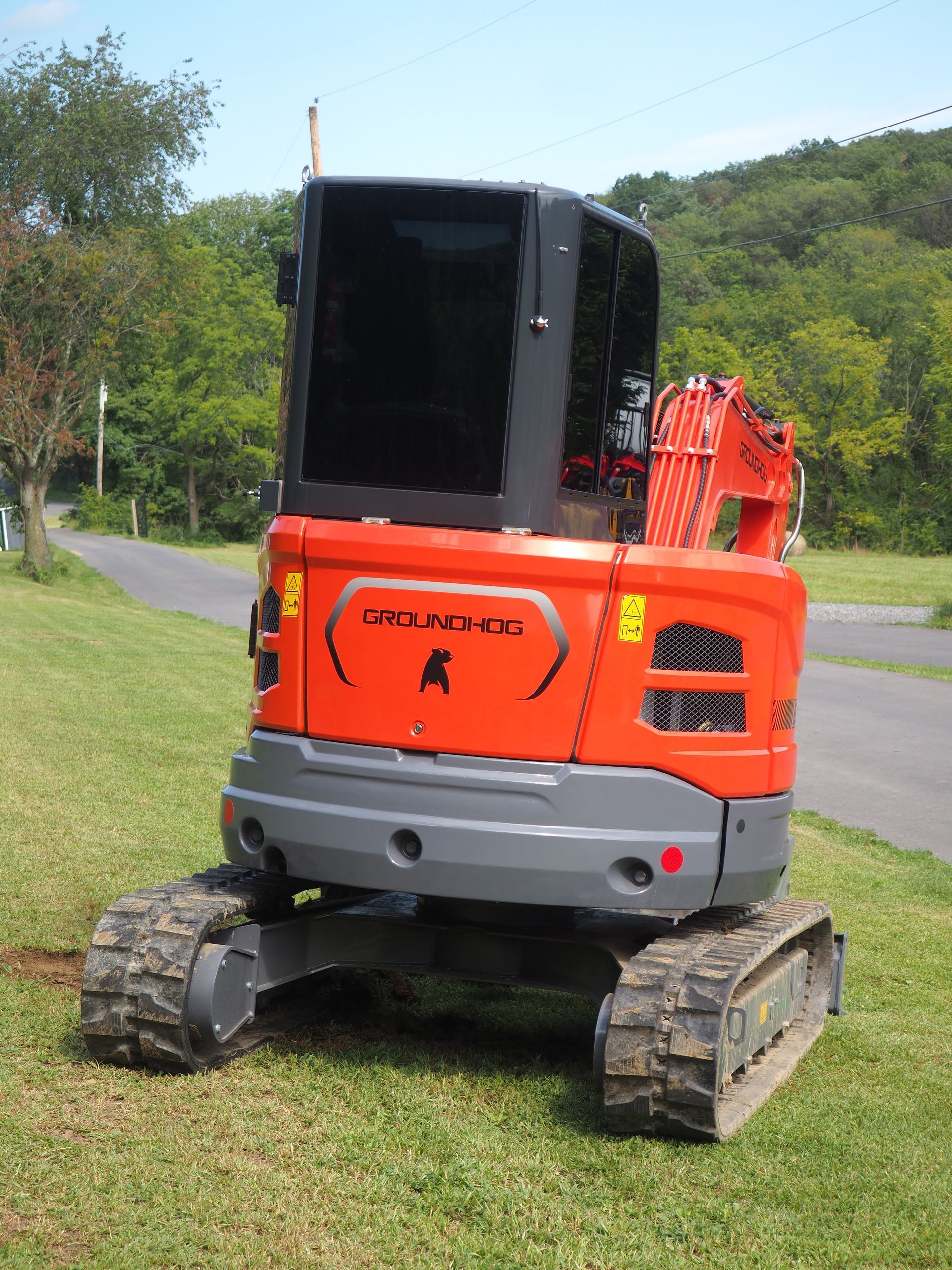 Orange Kubota excavator parked on grass. Black cabin, grey tracks, and arm are visible. Road and trees in the background.