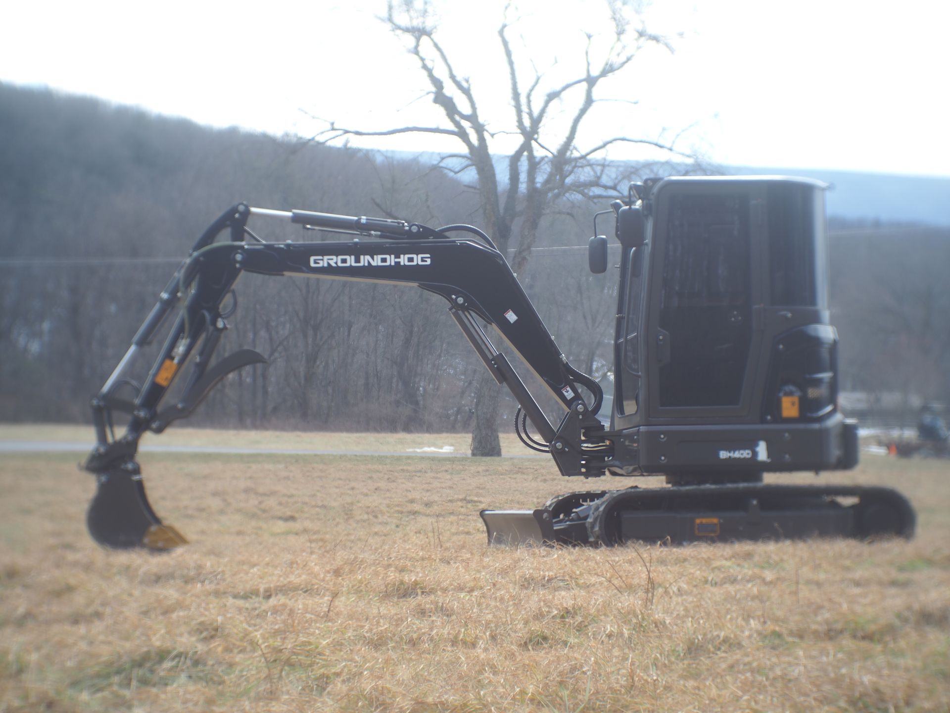 Black mini-excavator on a field, arm extended. Bare tree and hills in the background.