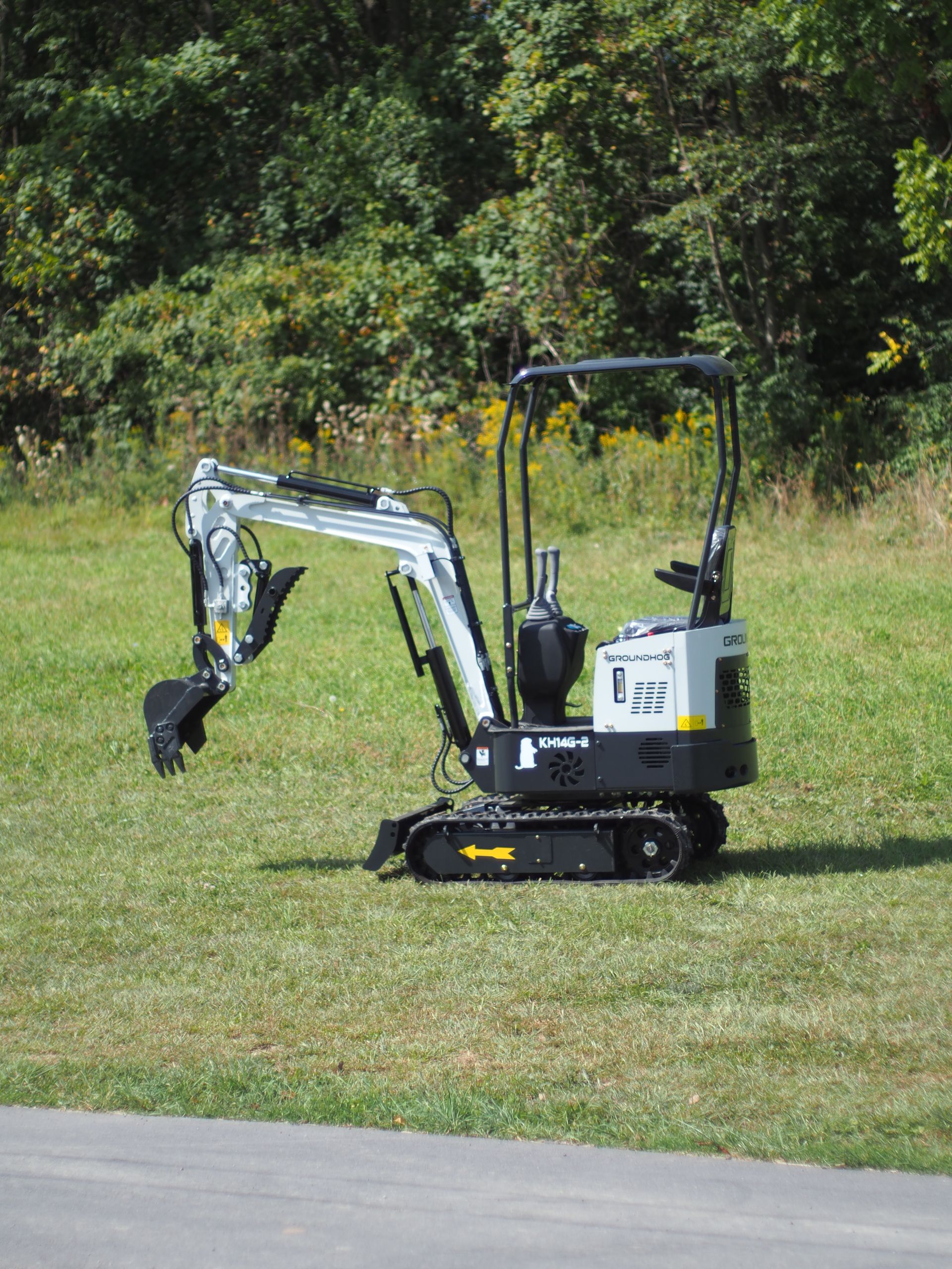 A small, gray and black excavator on green grass, with trees in the background.