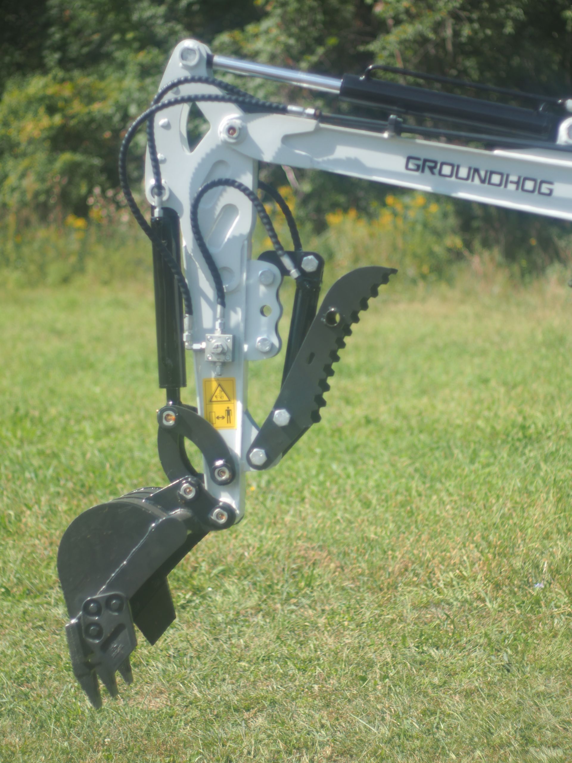 Groundhog mini-excavator arm with a black bucket and grapple attachment, set in a grassy field.