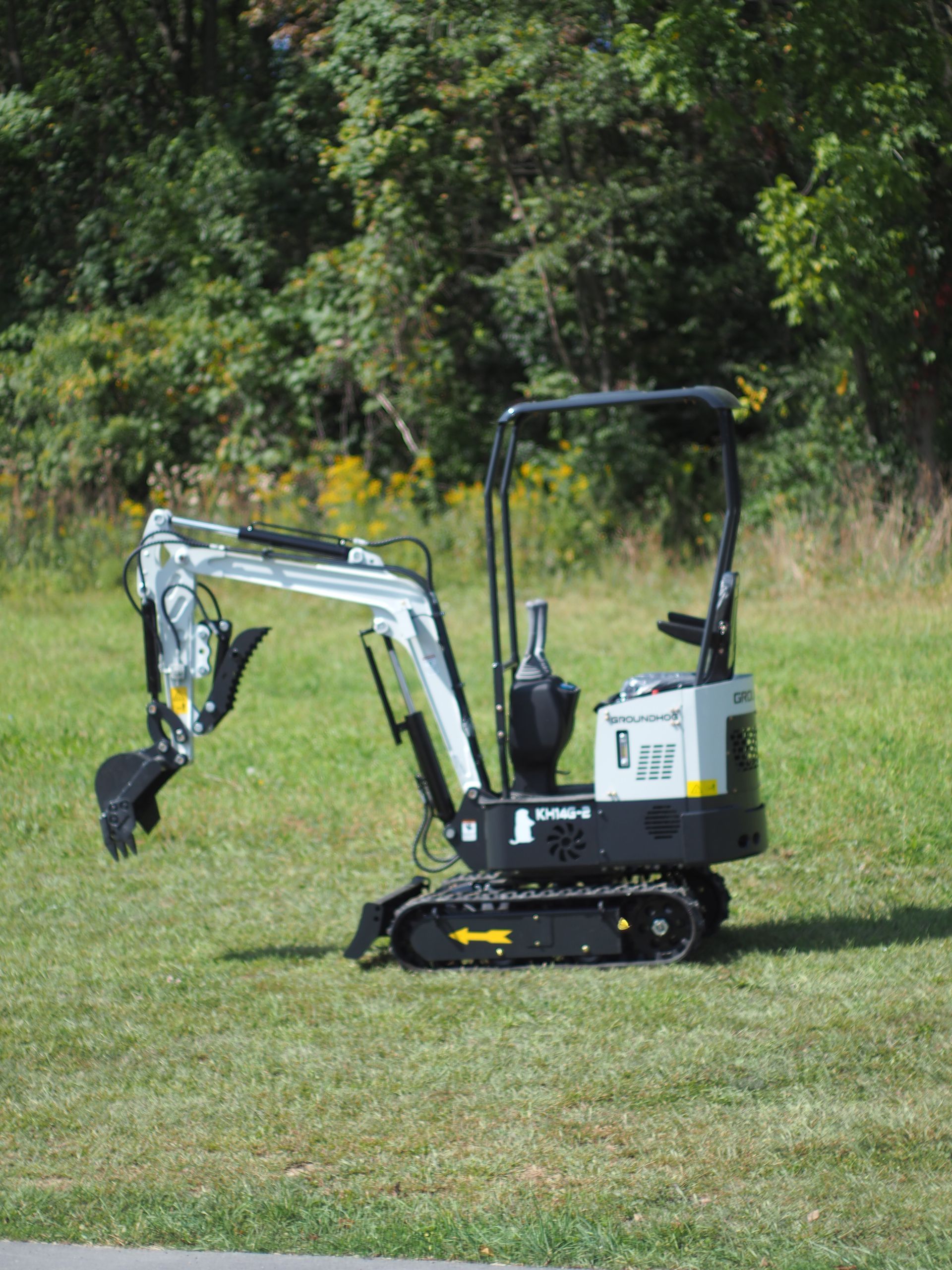 Mini excavator on grassy field, white and black, parked near trees on a sunny day.