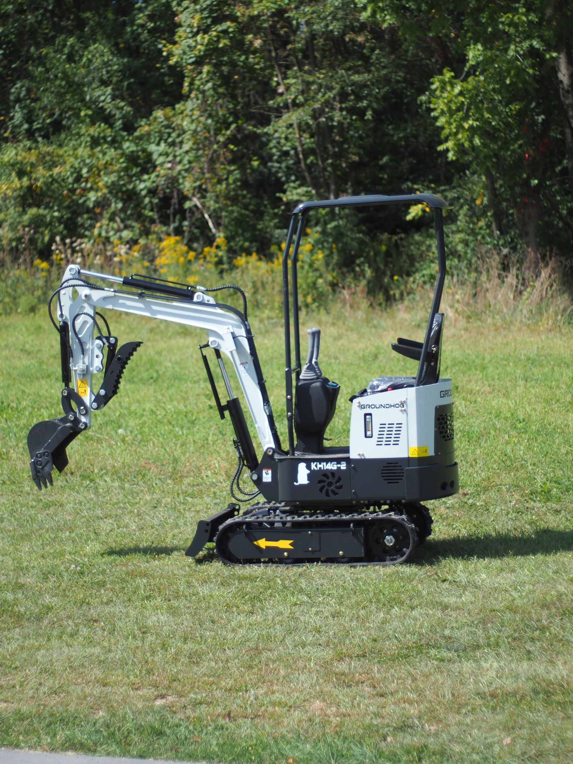 Mini excavator on grass, operating in daylight. White arm, black tracks and body, green trees in background.