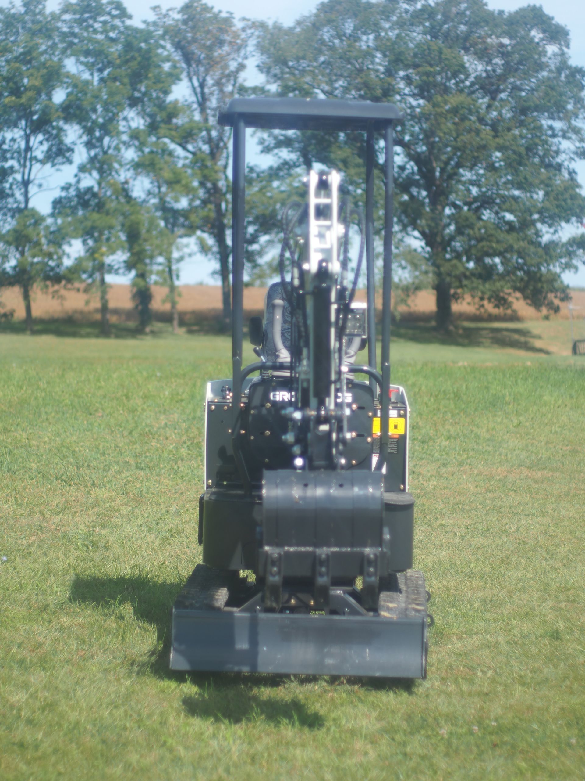Mini excavator on a grassy field. Black machine, protective cage, trees in the background.