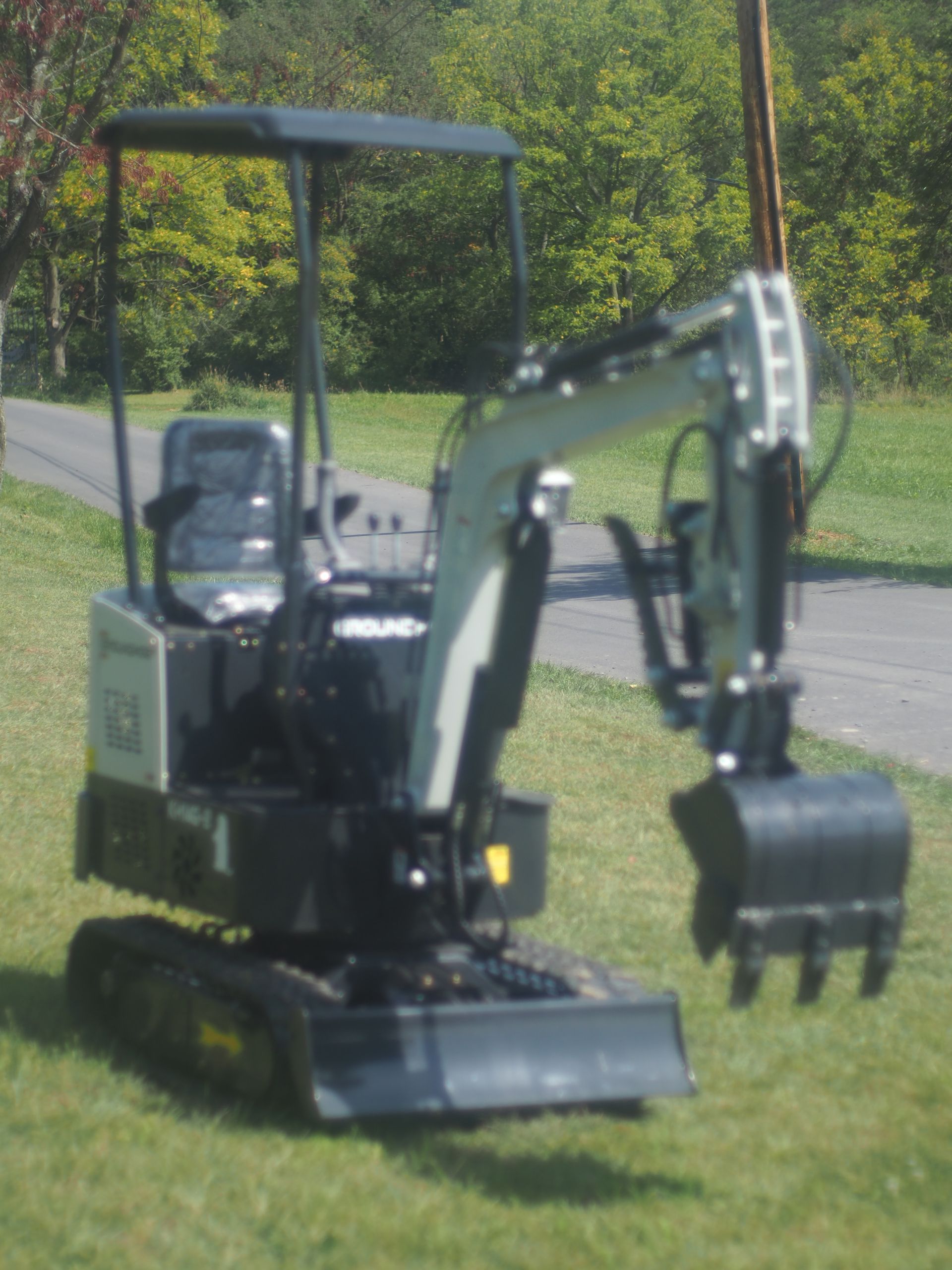 Mini excavator on grass. Black tracks, bucket, and body; gray arm; green trees in the background.