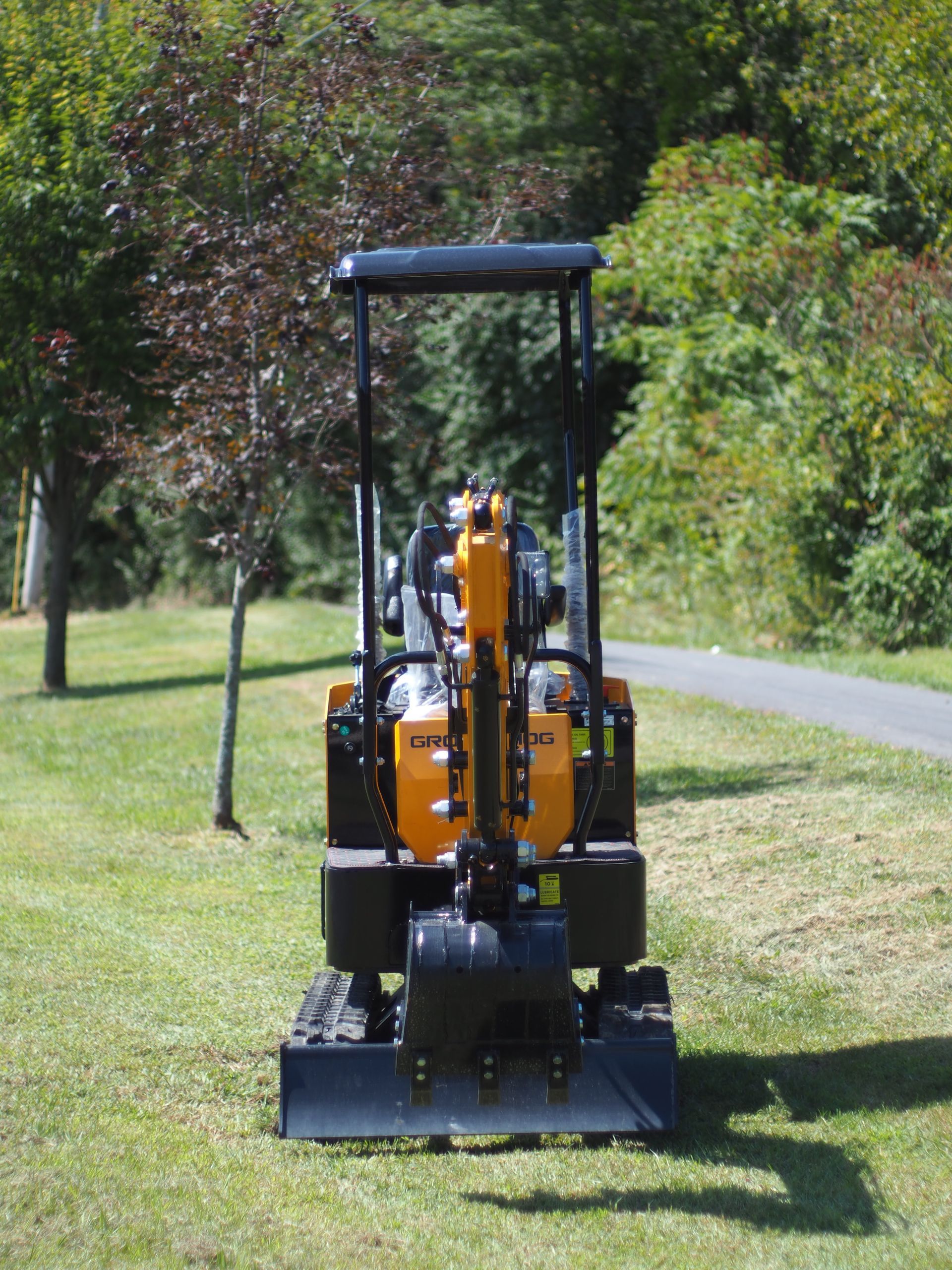 Mini excavator on grass, yellow and black, with trees and a path in the background.