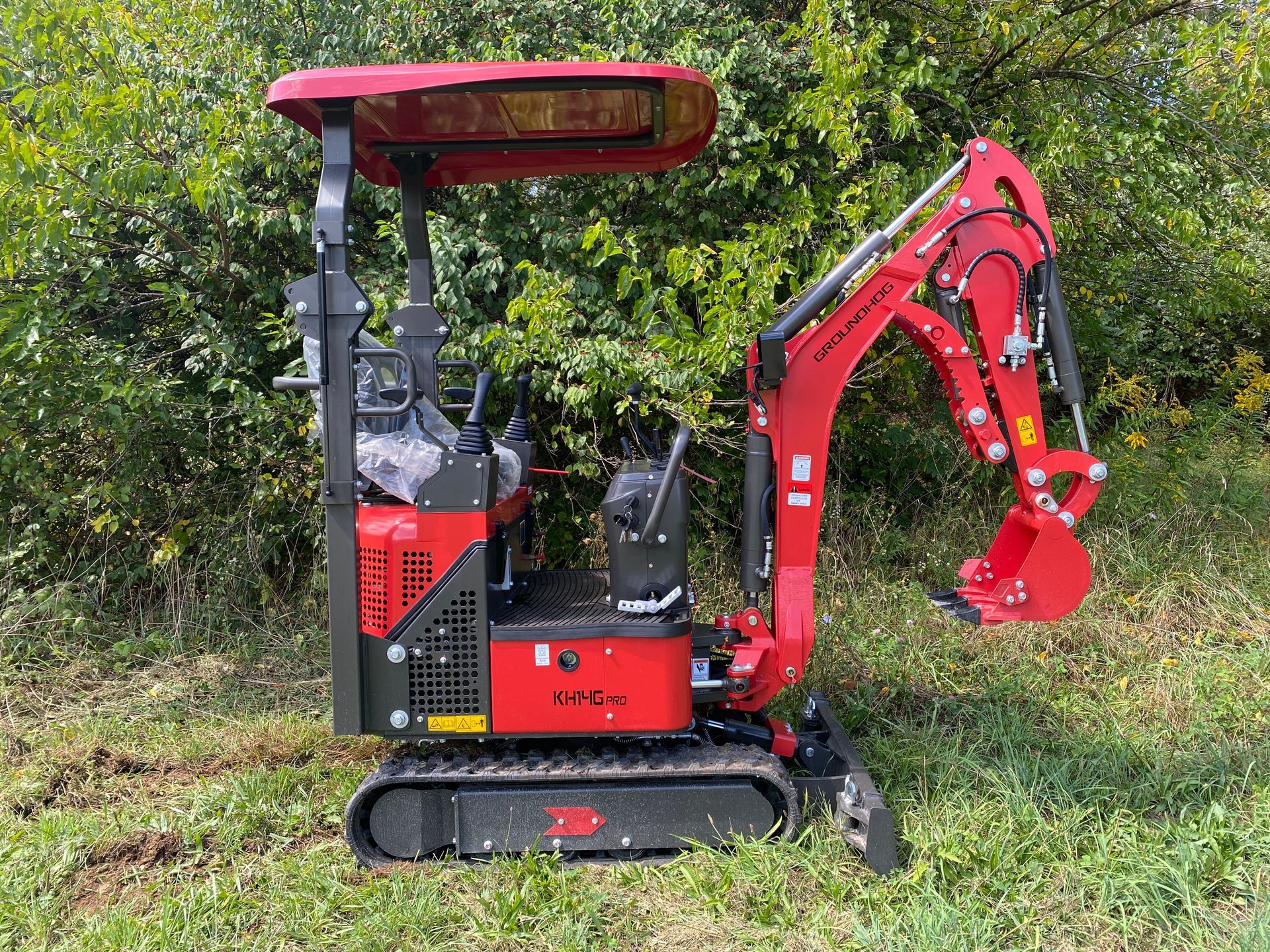 Red mini-excavator on grass with canopy, ready for digging; green foliage in the background.
