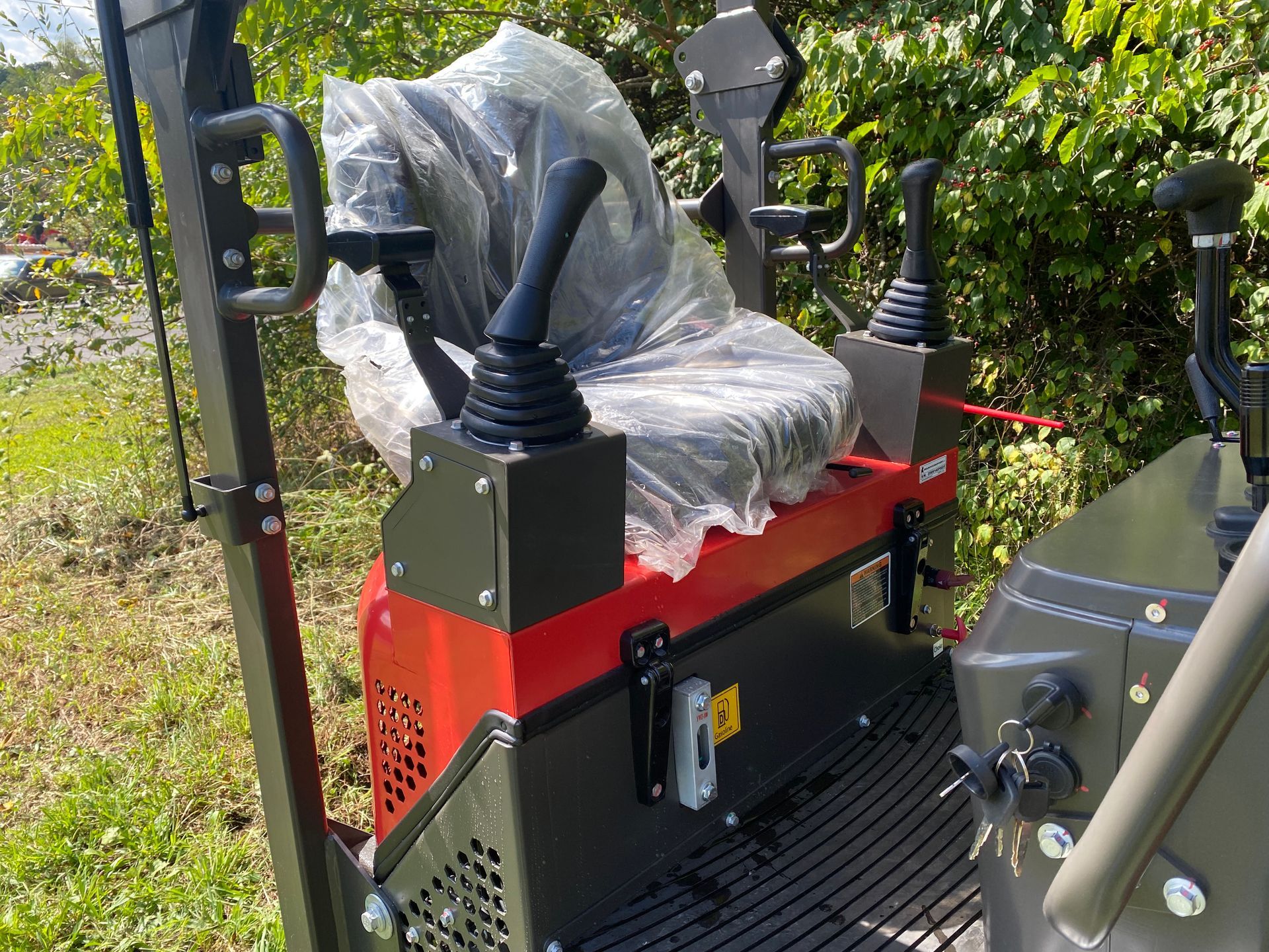 Mini excavator cab with seat, controls, and protective plastic wrap. Red and black, outdoors.