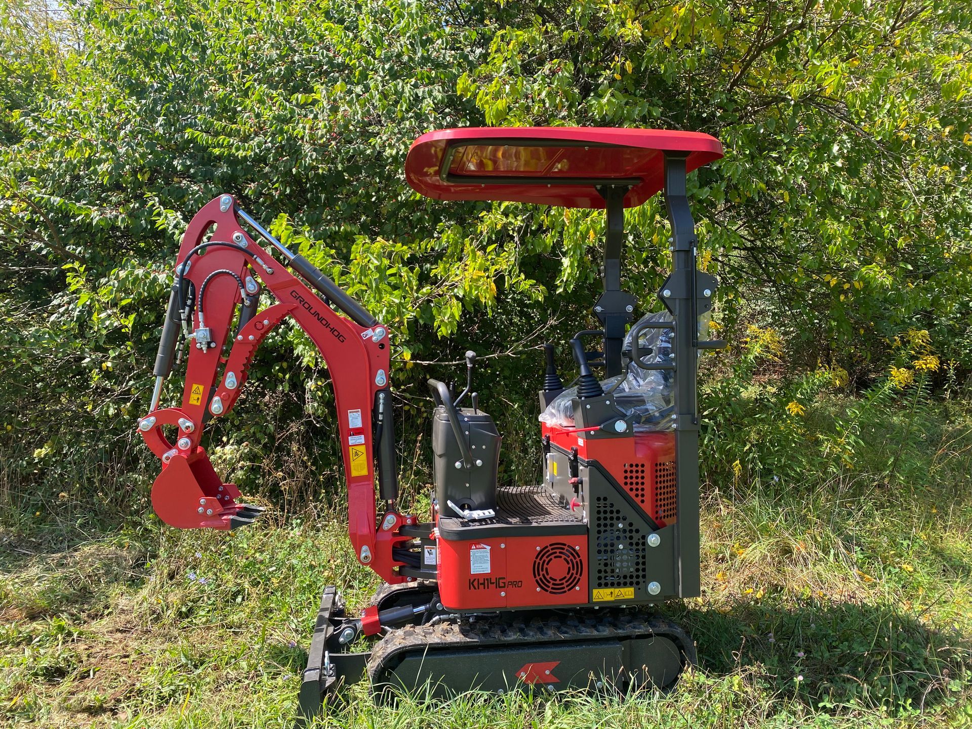Red mini excavator on tracks in a grassy area, with a canopy and foliage background.