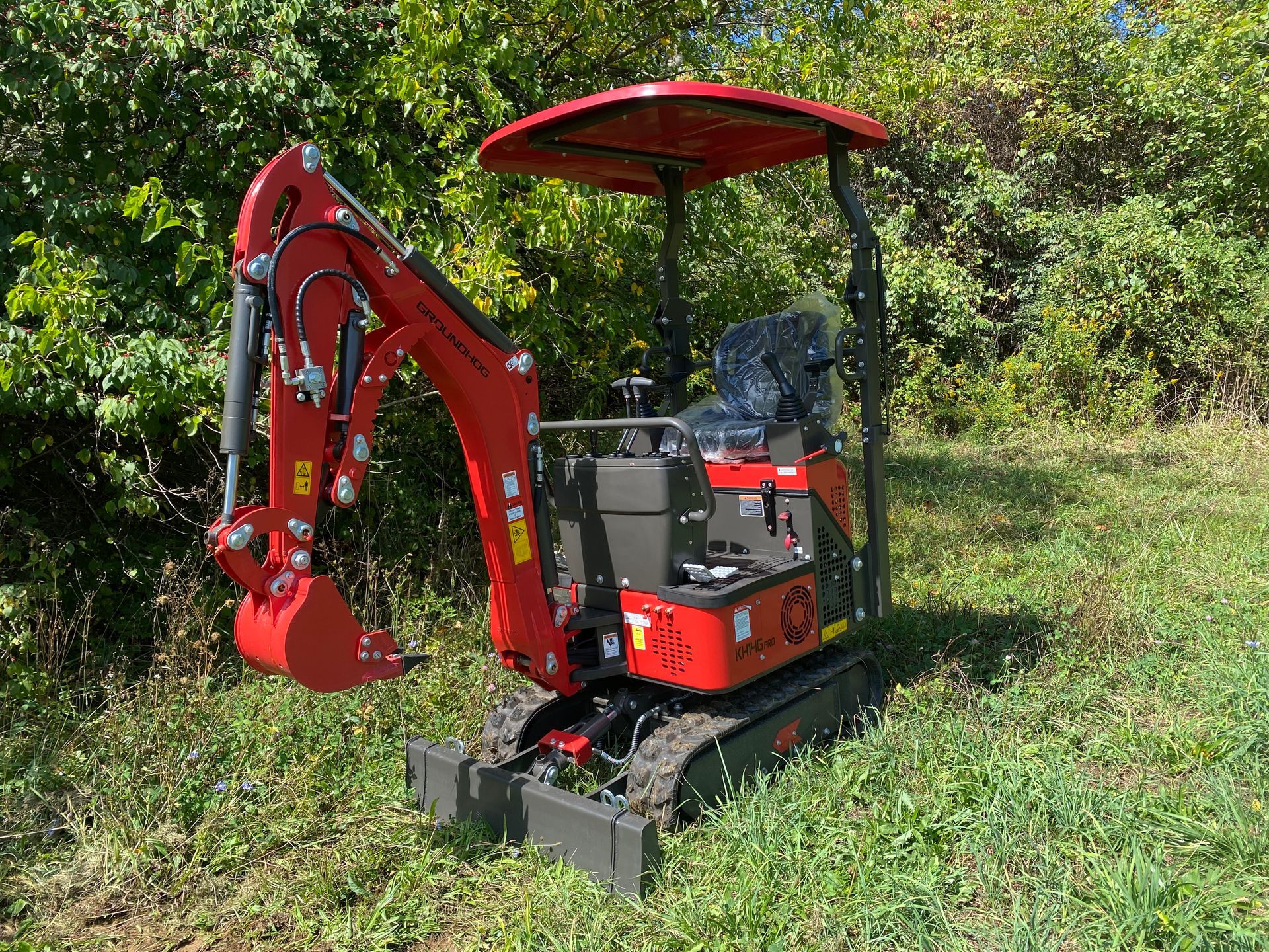 Red mini excavator on grassy ground with a canopy, in a wooded area.