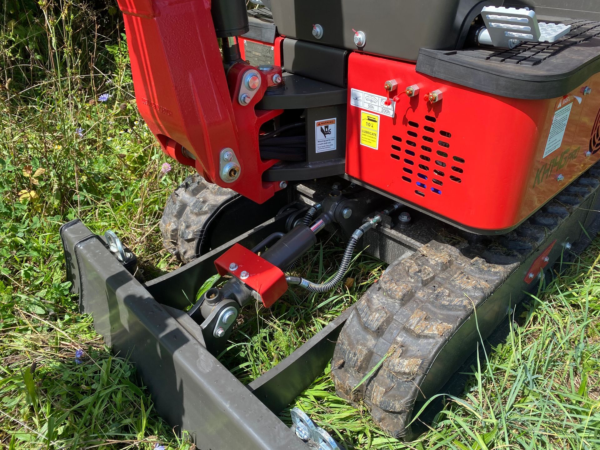 Red and black mini-excavator with tracks on grassy ground, showing blade, hydraulic system, and engine compartment.